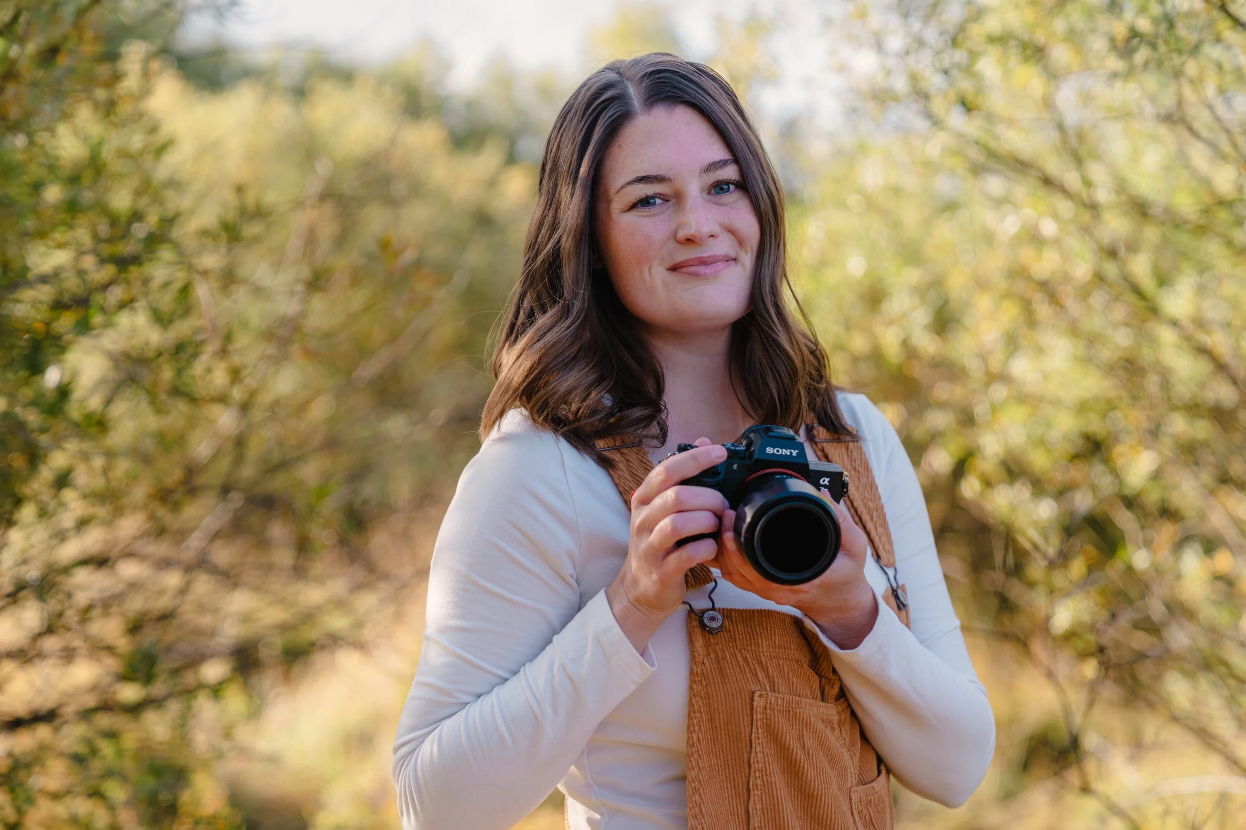 A woman with long brown hair and blue eyes holding a Sony camera, standing outdoors in a wooded area with yellow-green foliage.