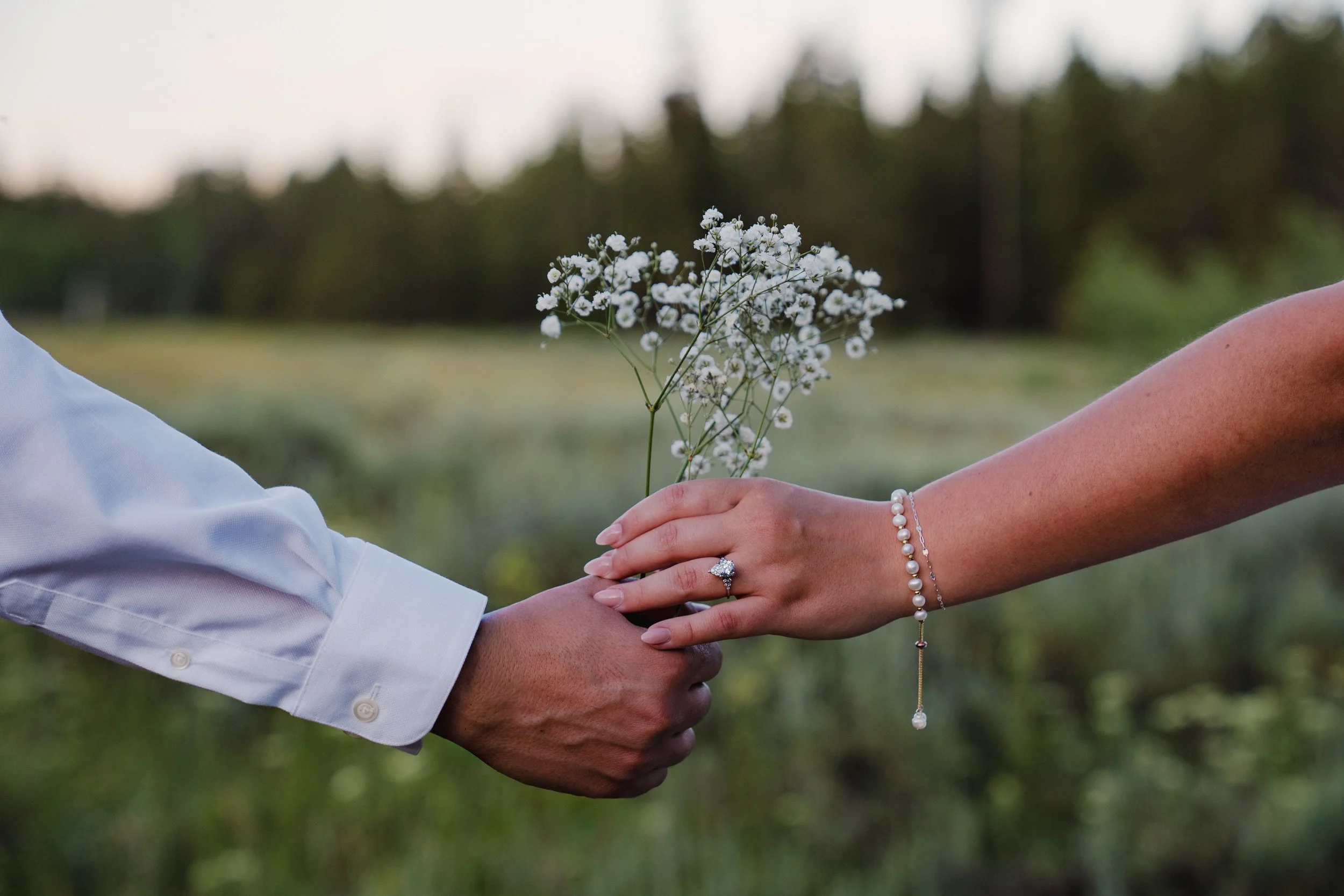 A couple holding hands outdoors, with the woman offering a bouquet of small white flowers to the man. The woman's hand is adorned with rings and pearl jewelry, and the man is dressed in a white shirt.
