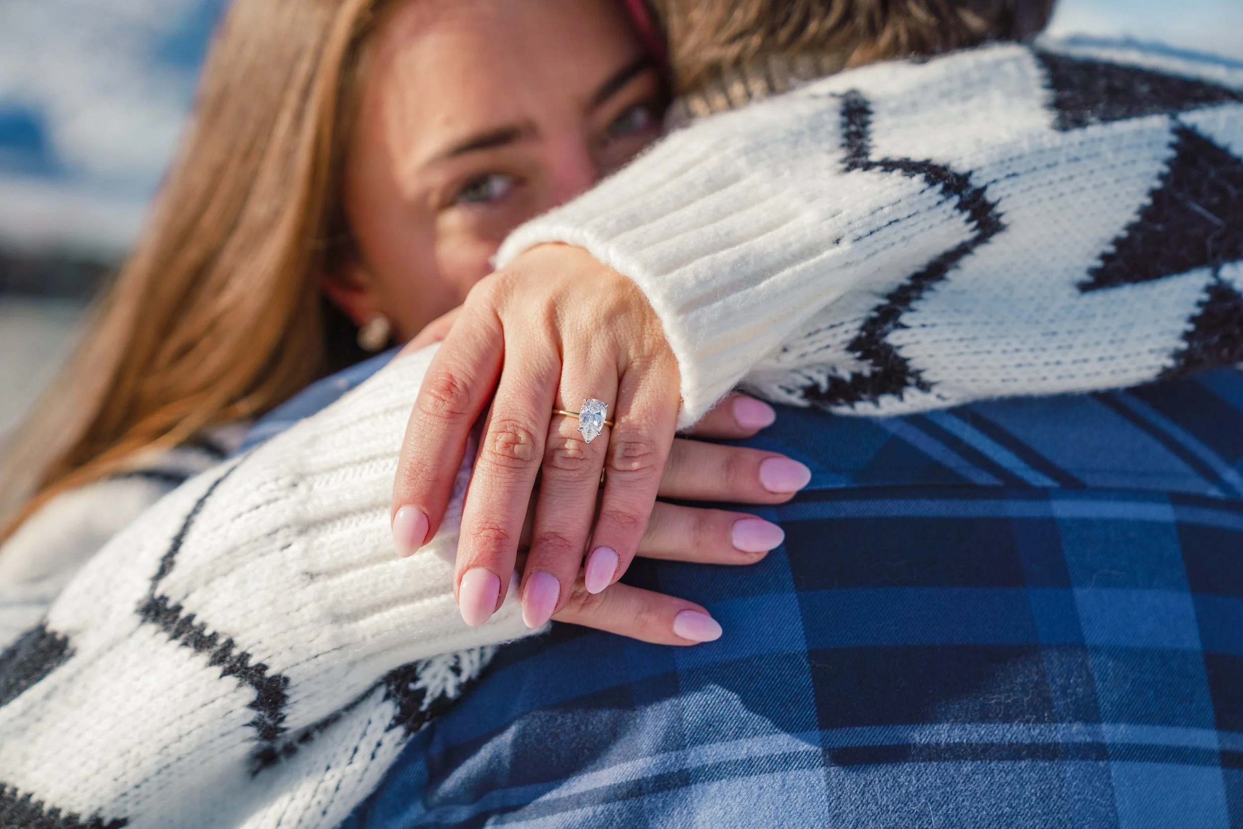 A woman with red hair and blue eyes embraces someone wearing a white sweater with black stripe patterns, showing an engagement ring with a large diamond on her finger.