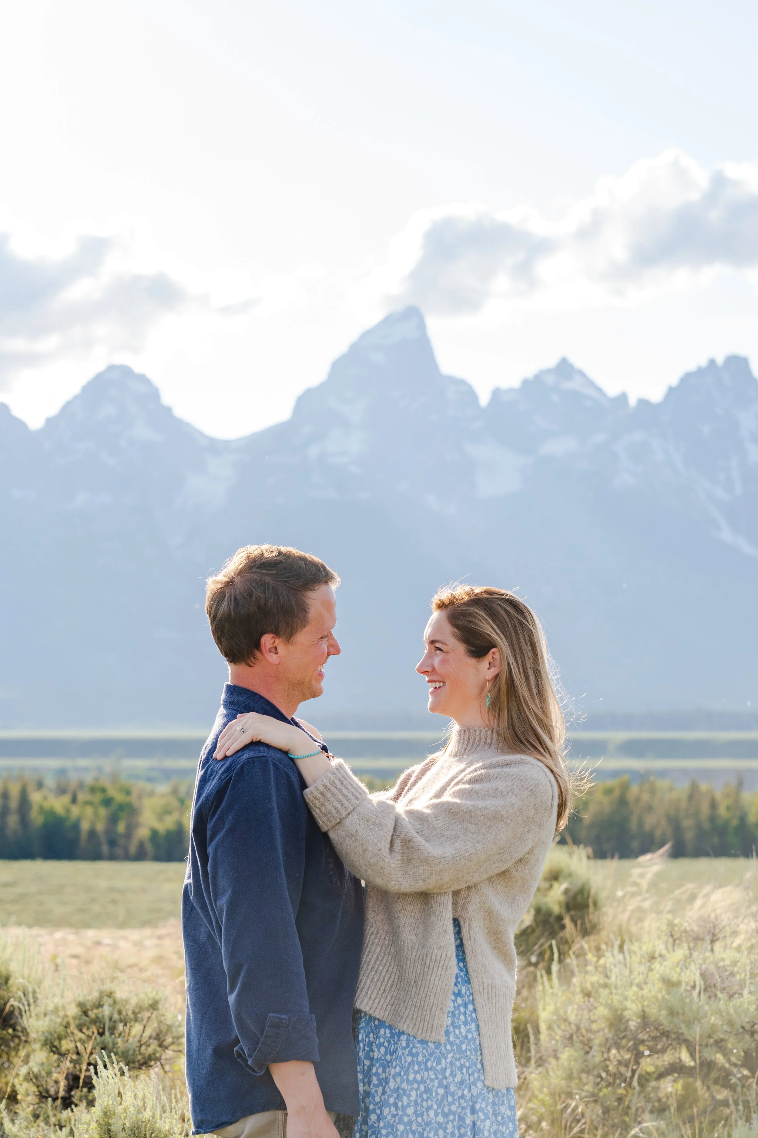 A couple standing outdoors, smiling and facing each other, with the Tetons and cloudy sky in the background.