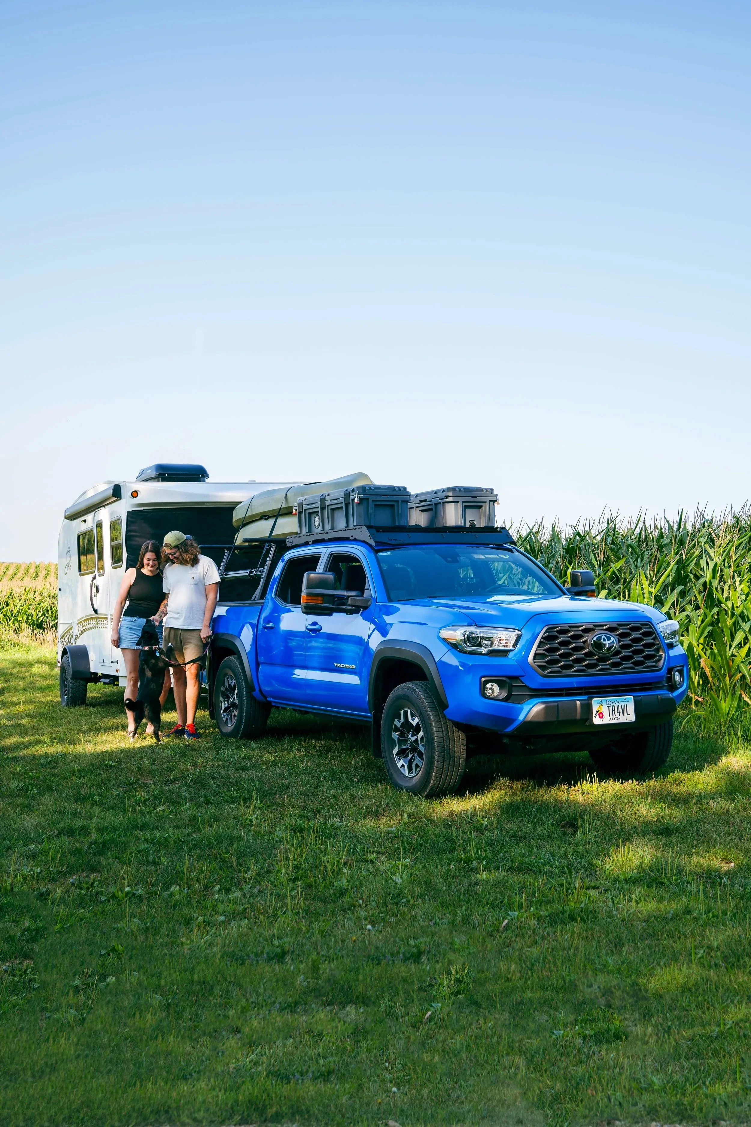 A blue Toyota Tacoma pickup truck parked on a grassy field next to a white travel trailer with two women and a dog standing beside the truck.