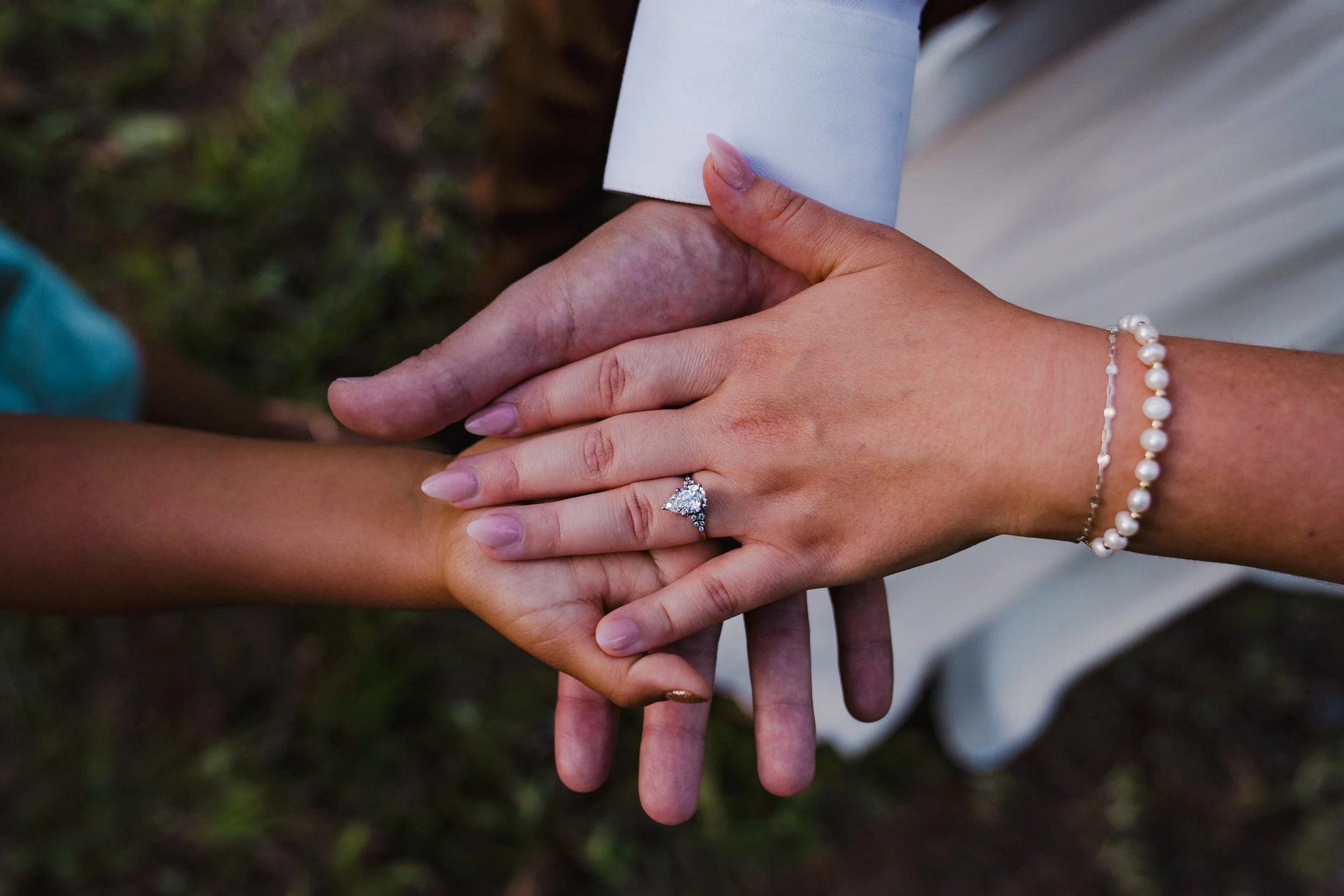 Close-up of hands holding each other, one with an engagement ring and pearl bracelet, the other with a wedding band, against a blurred outdoor background.