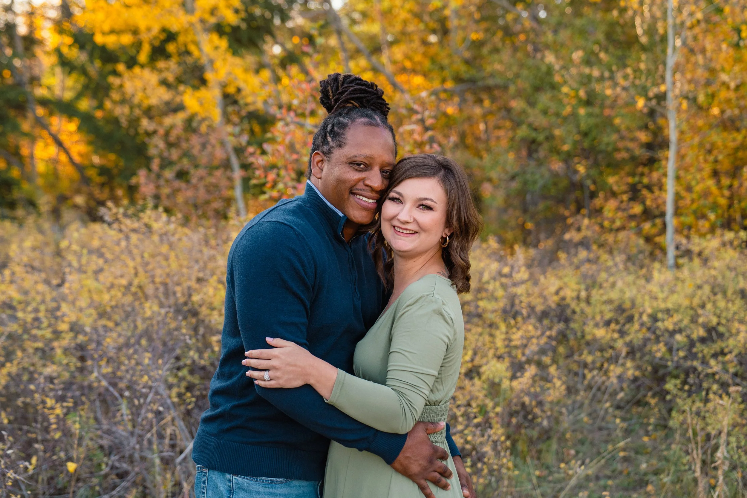A couple hugging outdoors during autumn with trees and fallen leaves in the background.