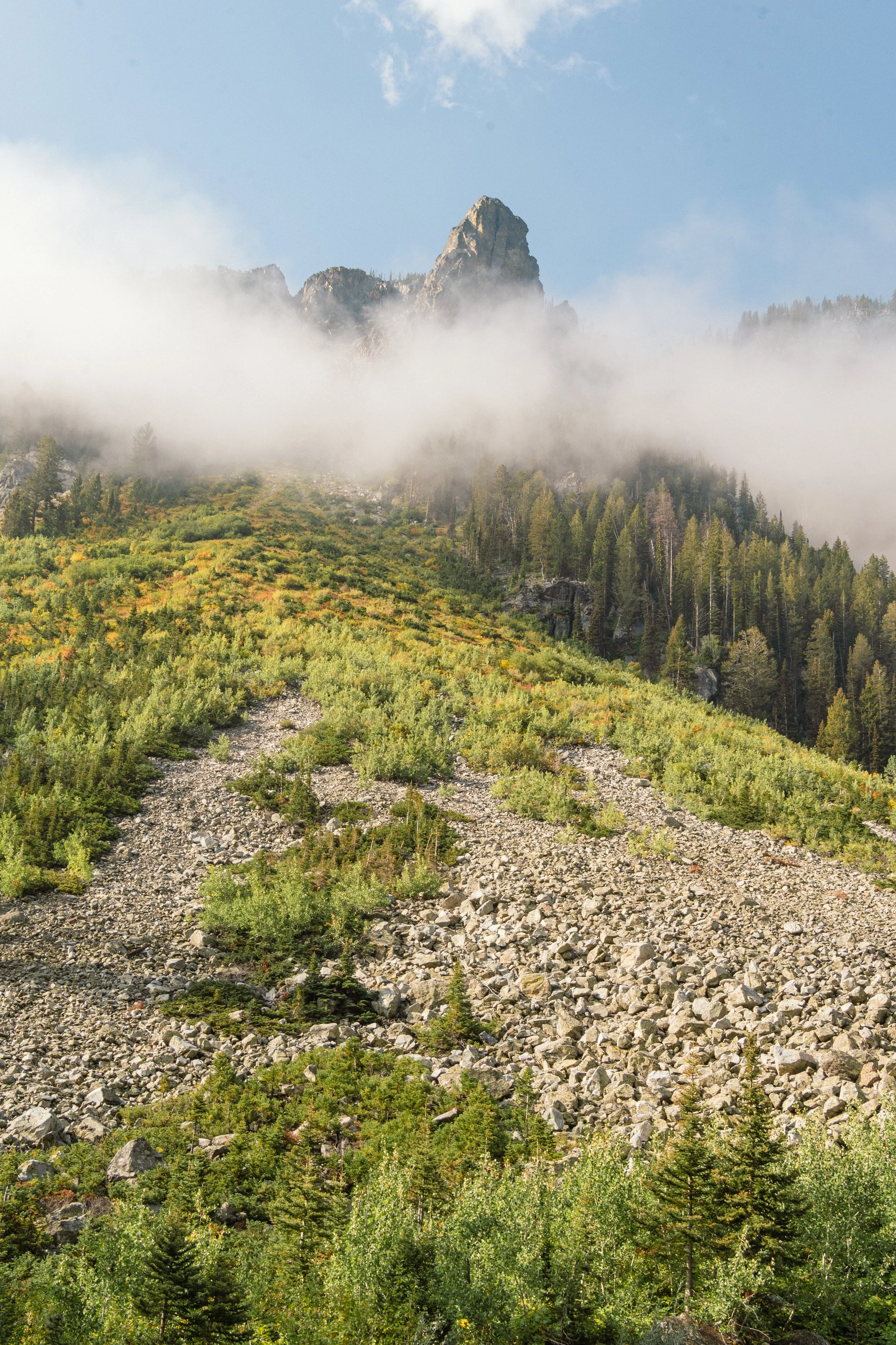 Mountain peak with fog and greenery below, rocky terrain in the foreground, and blue sky with clouds.