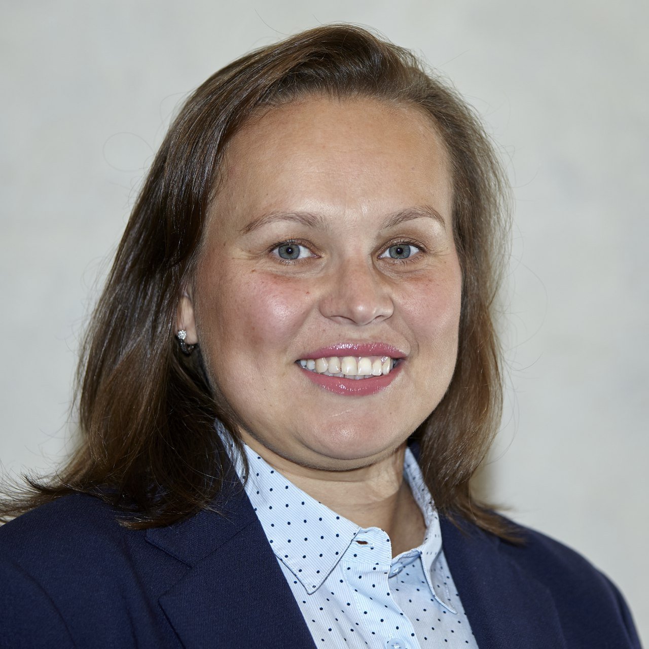 A woman with light skin, brown hair, wearing a dark blazer and a white shirt with tiny black dots, smiling.