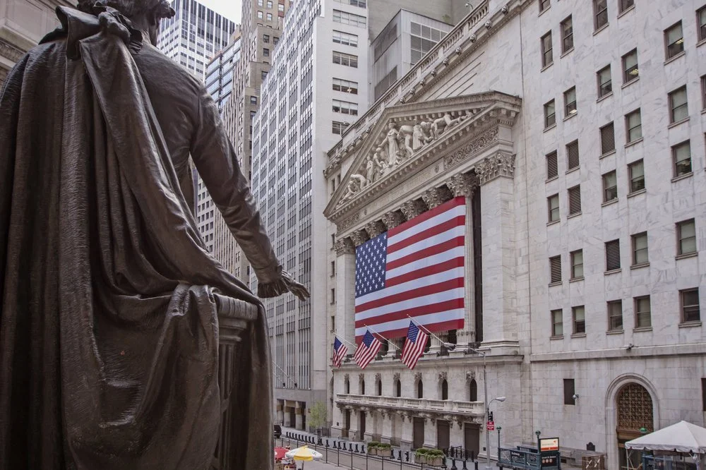 View of the New York Stock Exchange building adorned with the American flag and smaller flags, with a statue in the foreground.
