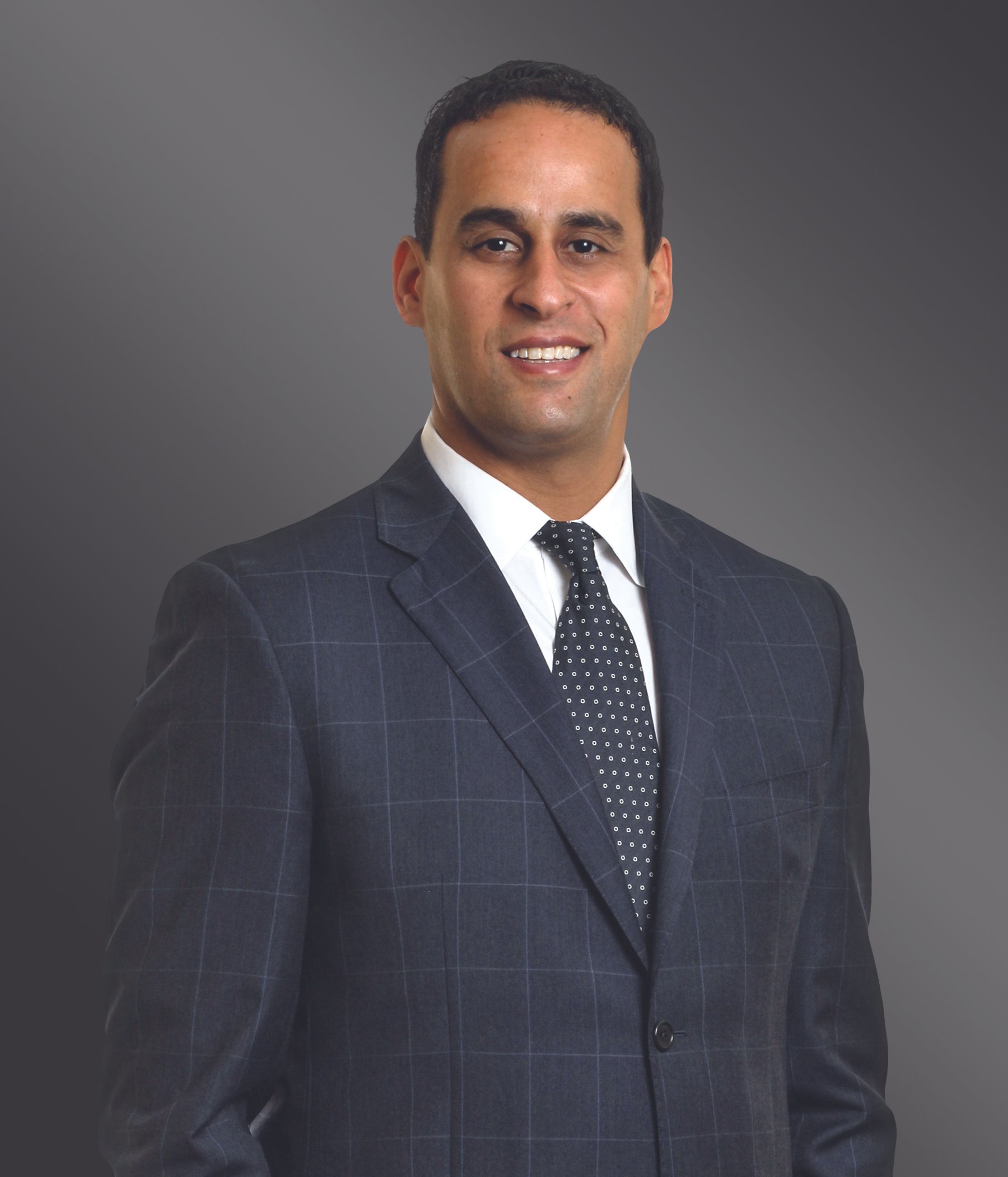 Professional man in dark plaid suit with white shirt and polka-dot tie, smiling, against a gray gradient background.
