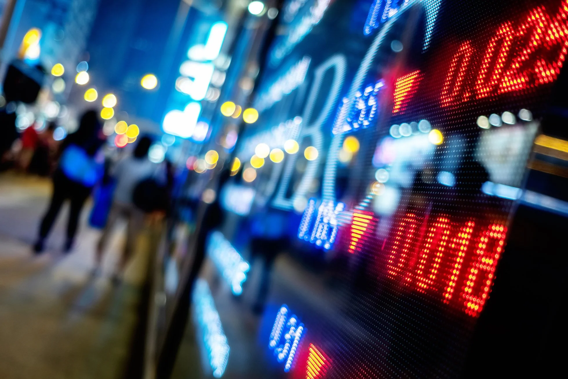 Blurred image of a digital stock market display with colorful blinking lights and numbers, with pedestrians walking by in the background at night.