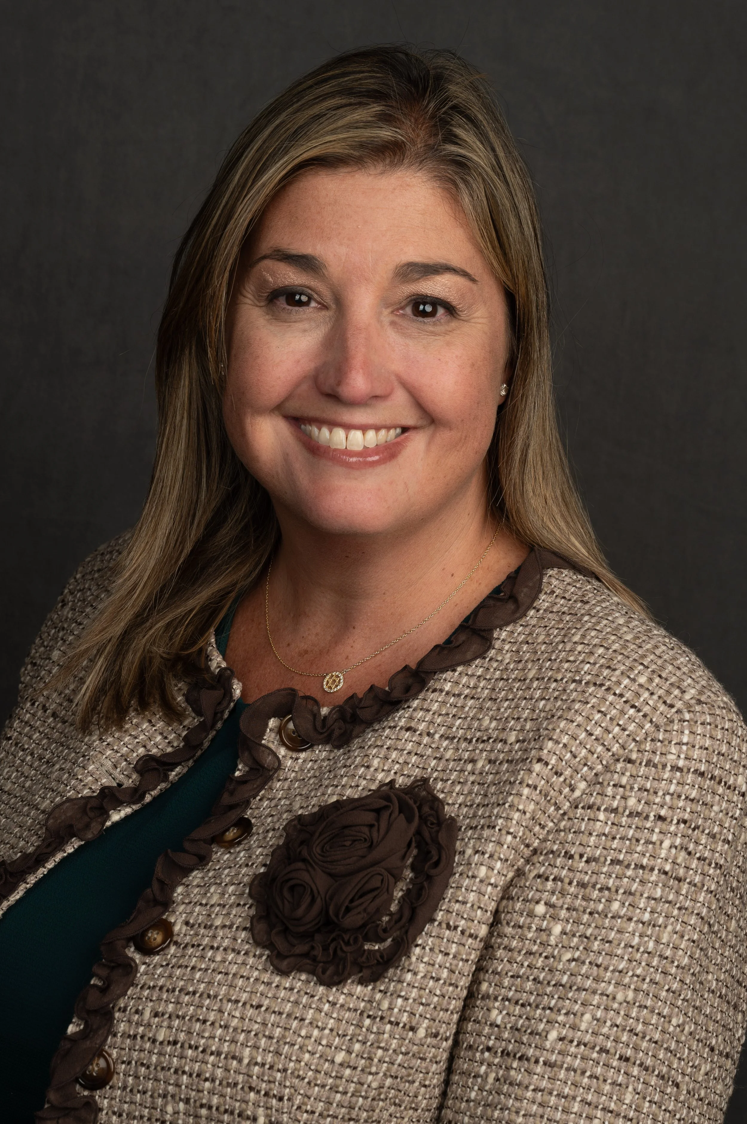 Portrait of a smiling woman with light brown hair, wearing a beige tweed blazer with brown ruffle trim and a large fabric flower, gold necklace, and pearl earrings, against a dark background.