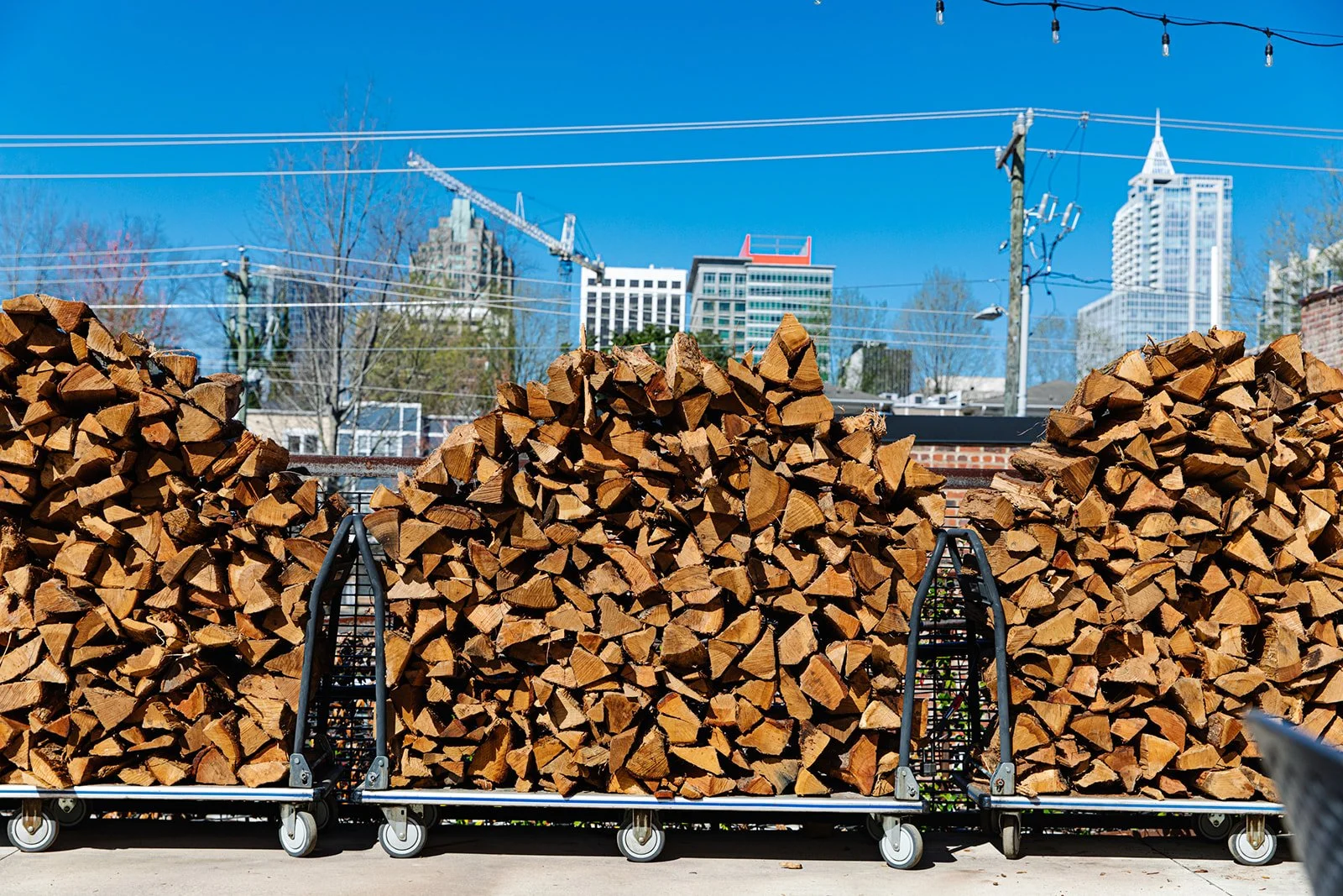 A large bundle of cut firewood stacked on a flatbed cart with wheels, set outdoors with the Raleigh city skyline in the background.