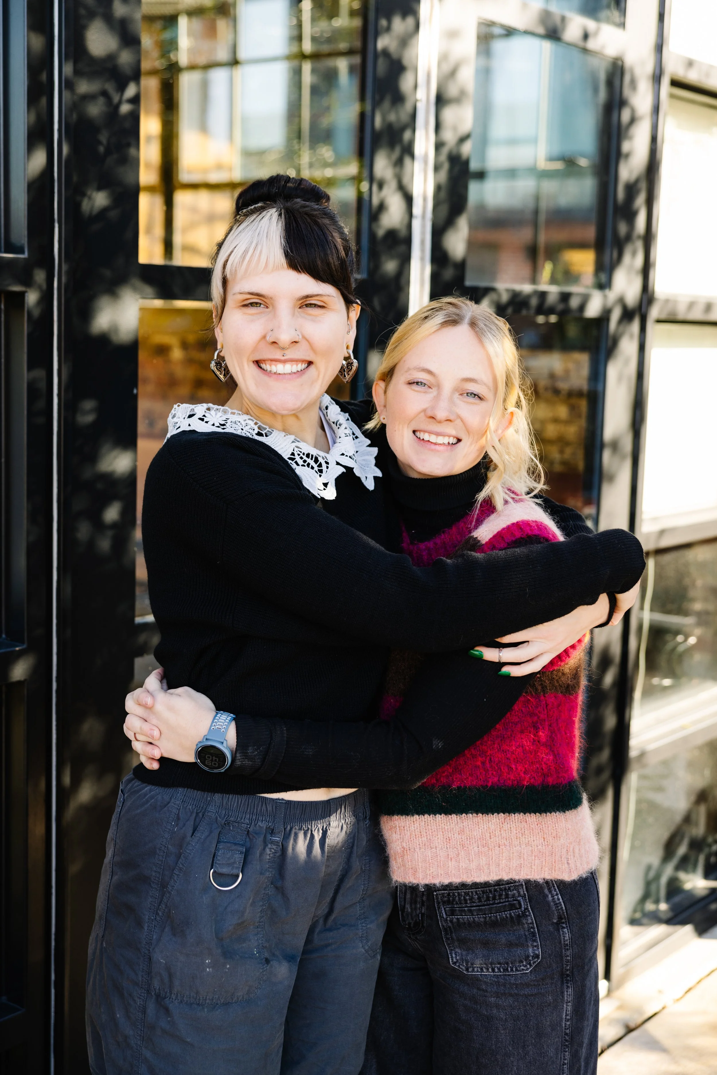 Two women hugging and smiling in front of a black metal and glass structure, with sunny outdoor setting.