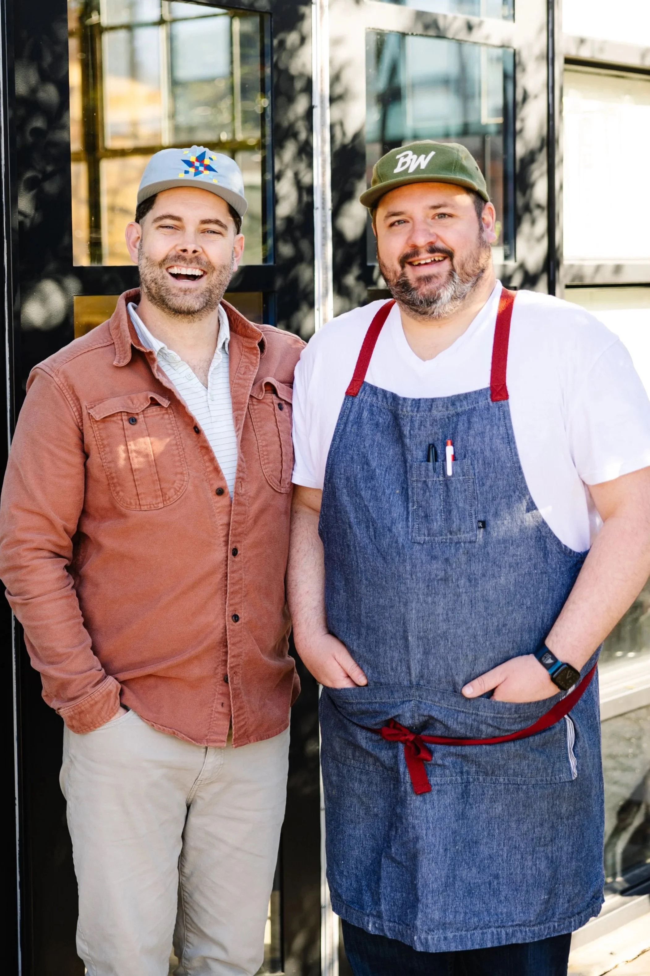 Two smiling men standing outside  modern building; one wearing a light gray cap with a colorful geometric design, a rust-colored jacket, and light khaki pants; the other in a green cap with 'BW' embroidered, a white shirt, and a denim apron with red straps and a pen in the pocket.