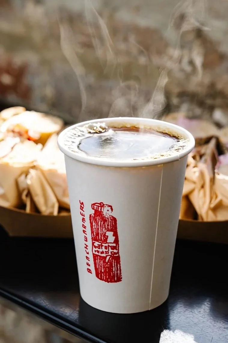 A hot cup of coffee in a white paper cup with a red logo, steam rising from the top, placed on a black surface with a tray of sandwiches in the background.