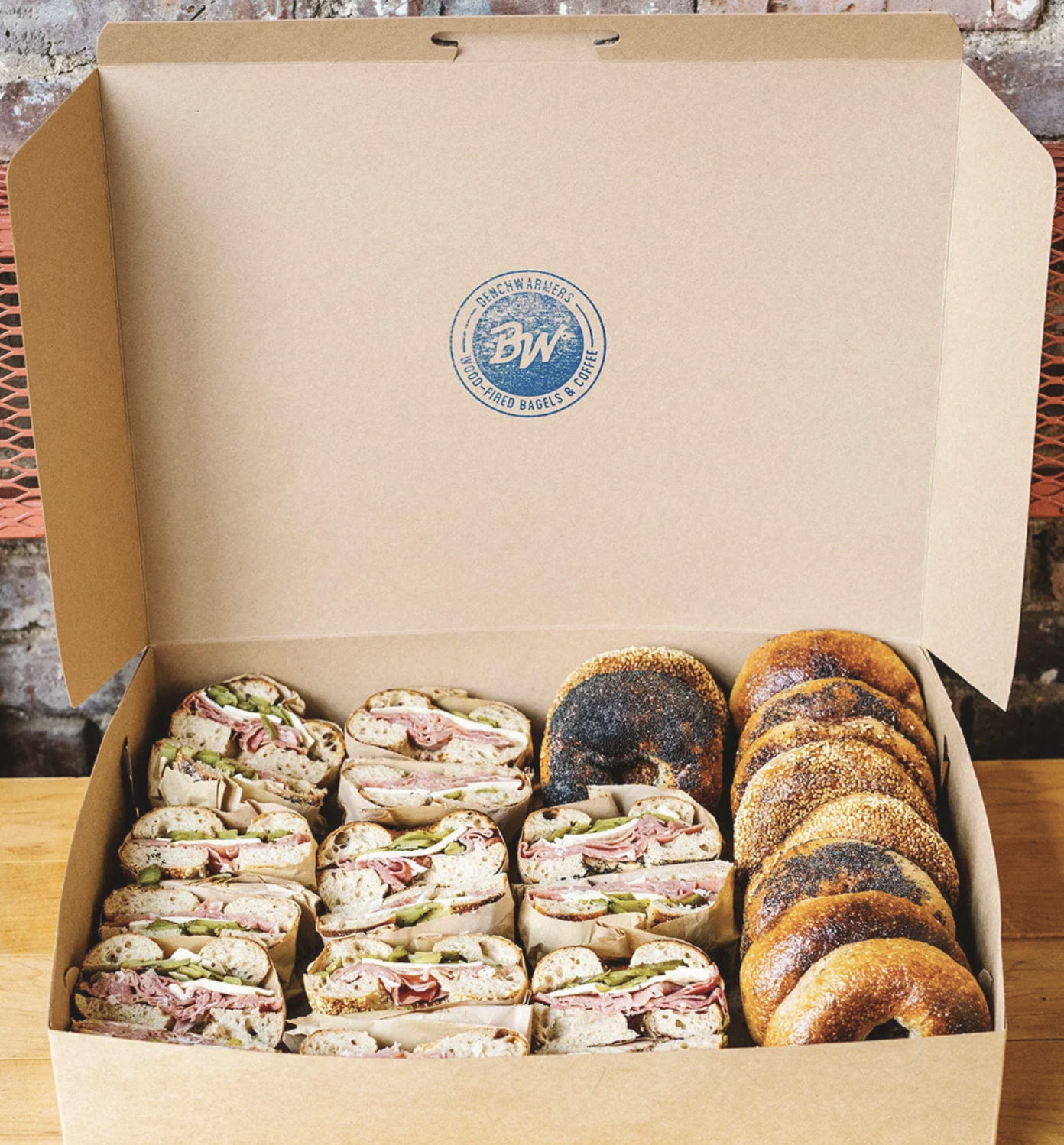 An open catering box containing multiple bagel sandwiches and bagels. The sandwiches are filled with meats, lettuce, and pickles, while the bagels are toasted and seasoned.