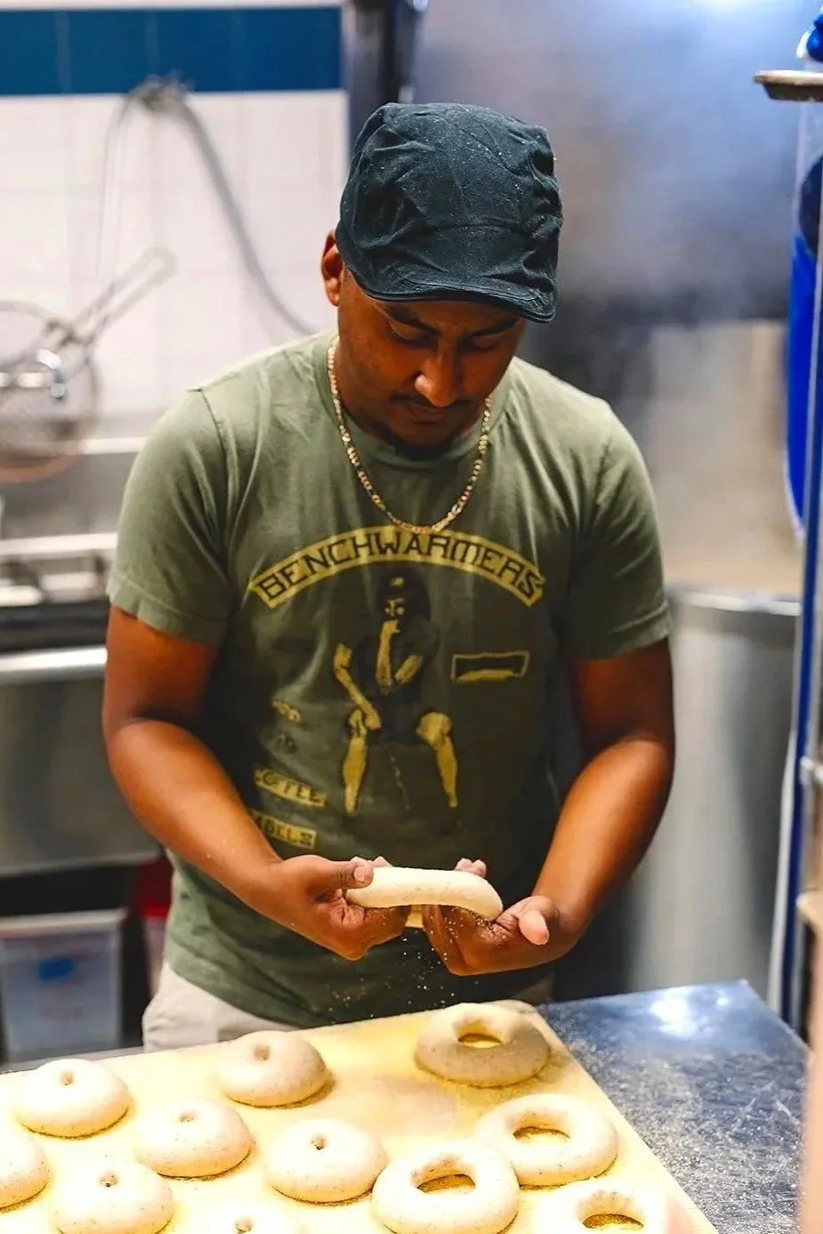 A man wearing a dark hat and a green Benchwarmers t-shirt is shaping bagel dough and preparing bagels on a wooden surface in our kitchen.