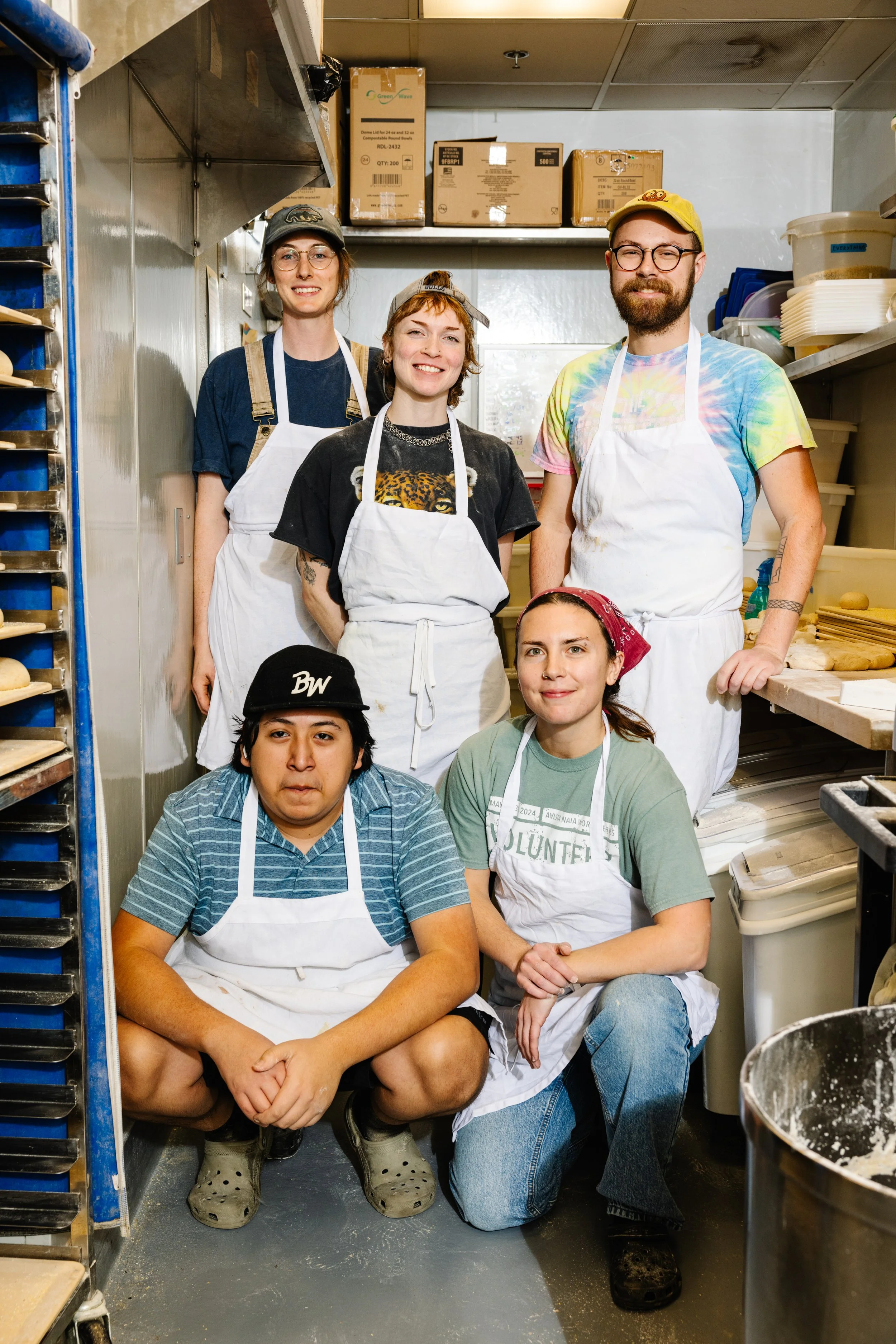 A group of five Benchwarmers bakers wearing aprons and casual clothing in our commercial kitchen. Three people are standing at the back, and two are kneeling int he front, surrounded by kitchen shelves and containers.