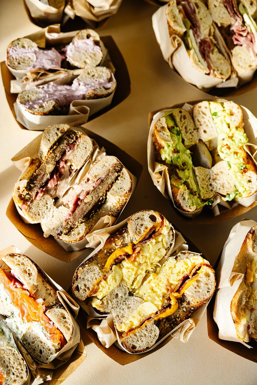 Various bagel sandwiches cut in half and arranged in paper-lined baskets, filled with ingredients like eggs, cheese, meats, lox, roe, veggies, and spreads.