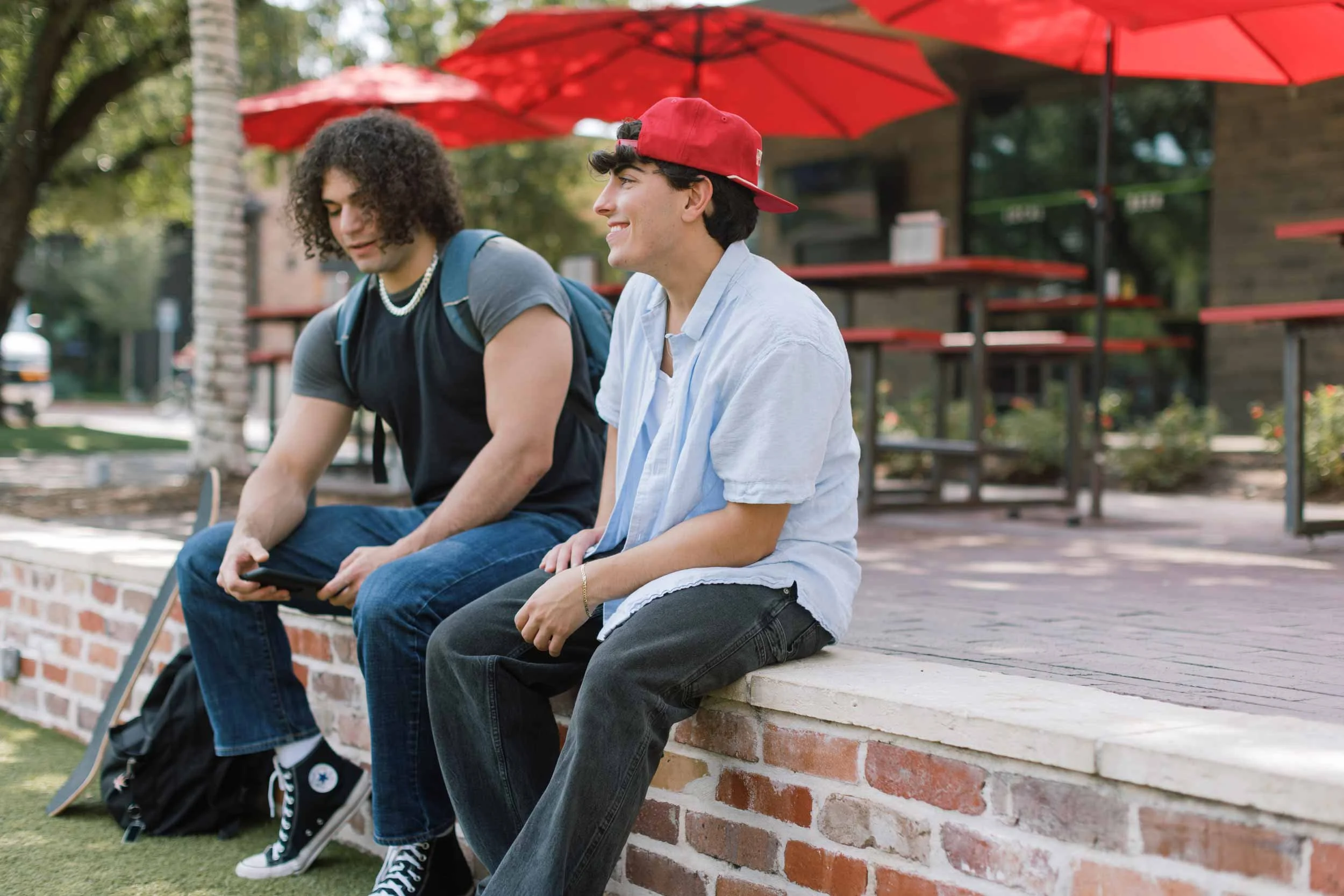 Two young men sitting on a brick ledge outdoors under red umbrellas, one using a phone, the other smiling, with trees and buildings in the background.