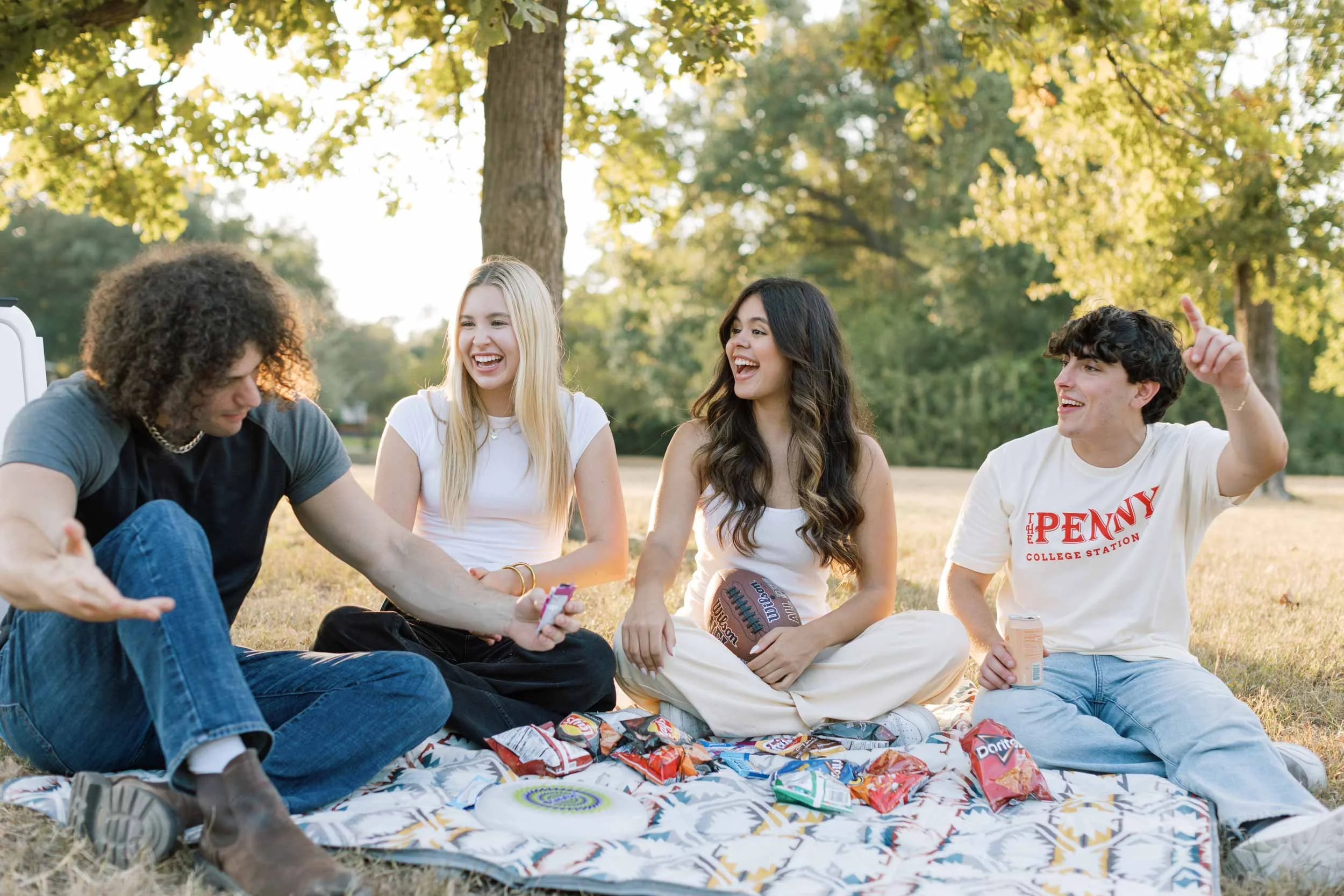 Group of five young friends sitting on a blanket outdoors in a park, laughing and talking, with snacks around them and a football.