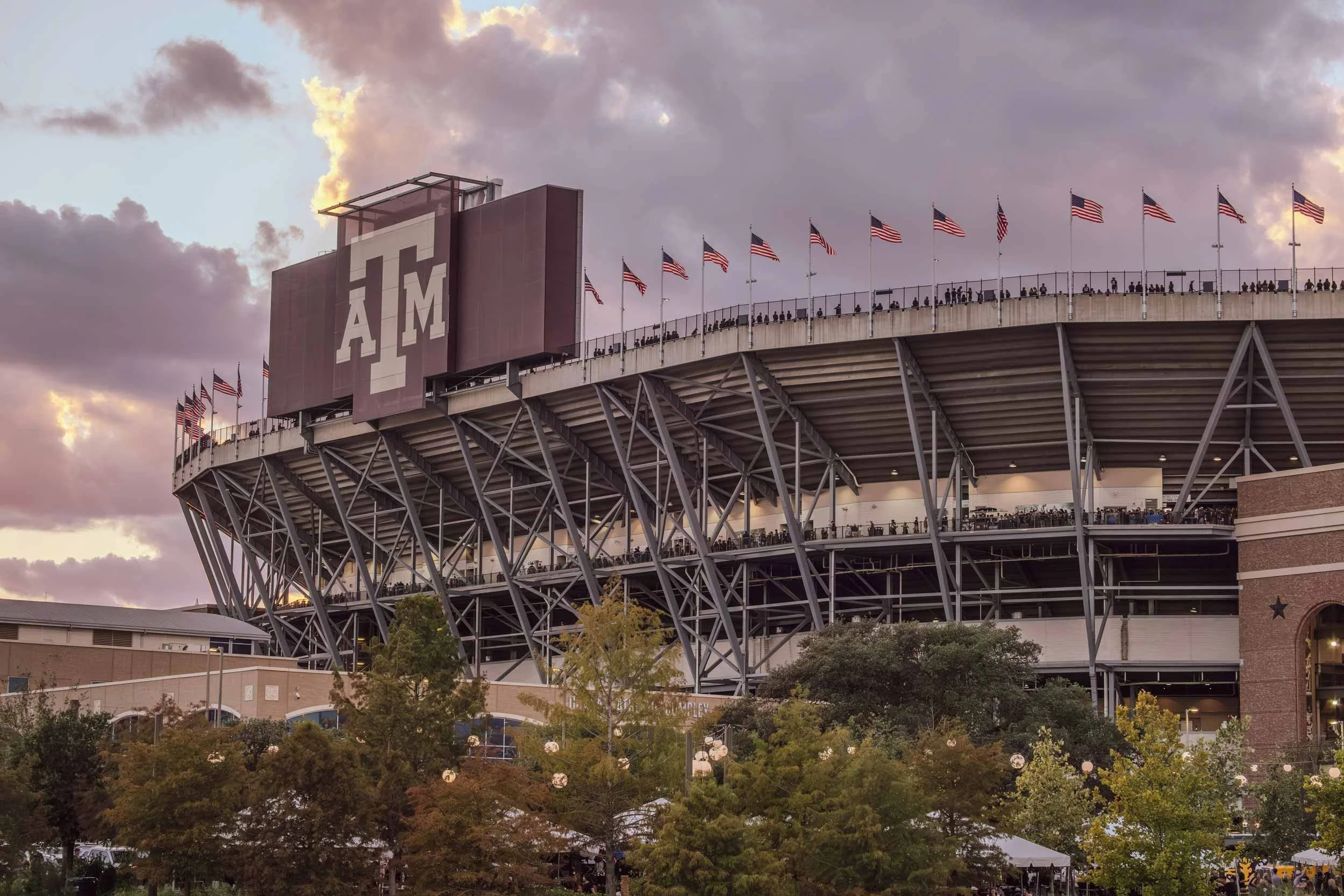 A large stadium with the Texas A&M University logo on a sign. The stadium is decorated with American flags along the top edge, and there are people seated in the stands. The sky is cloudy with a hint of sunset or sunrise light.