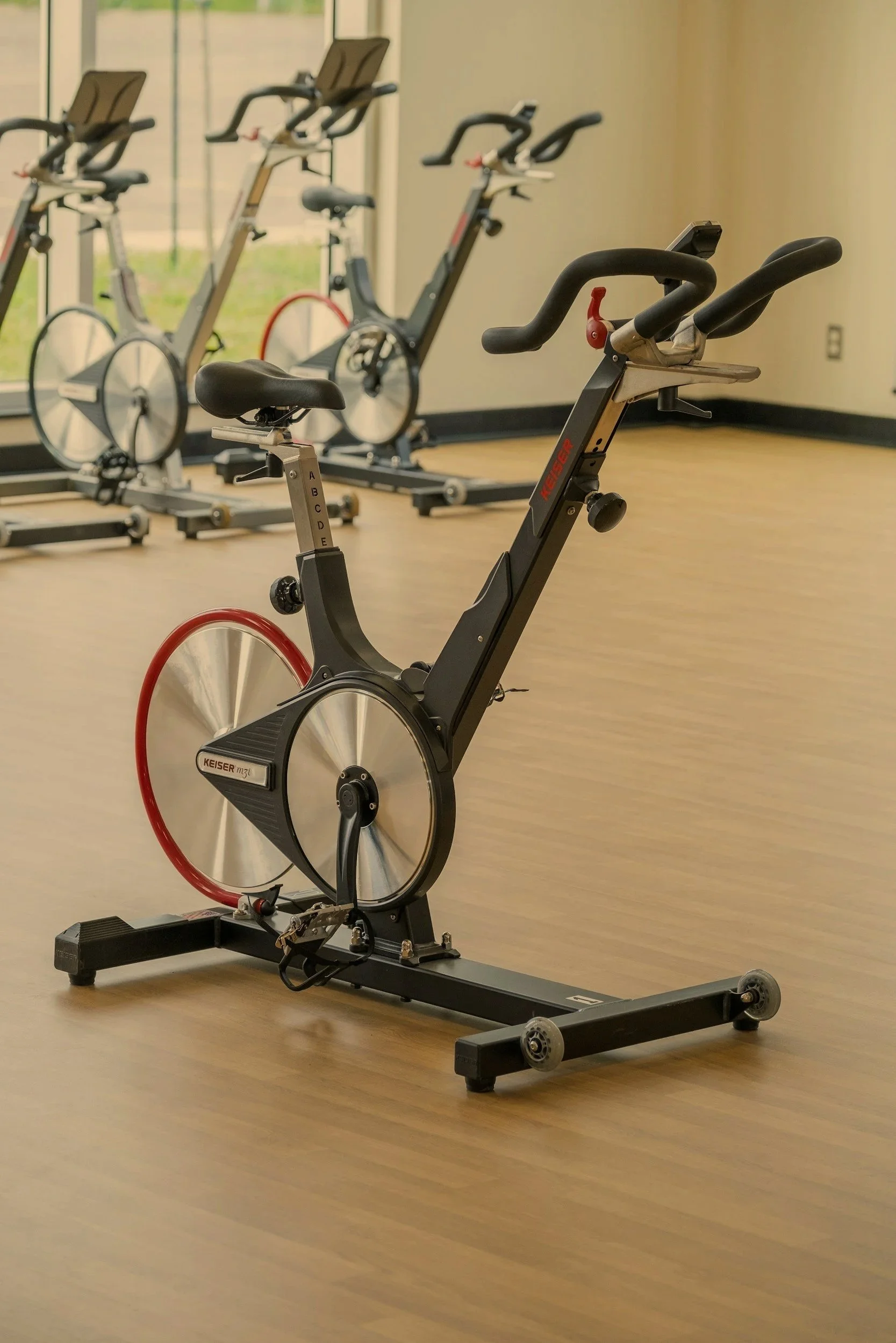 Indoor gym with multiple exercise bikes lined up on a wooden floor, large windows in the background allowing natural light.