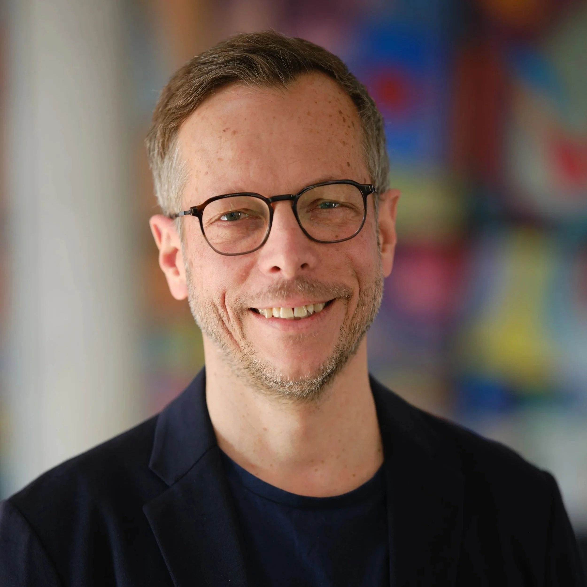 Close-up of a smiling man with glasses and freckles, wearing a dark suit, with colorful blurred background.