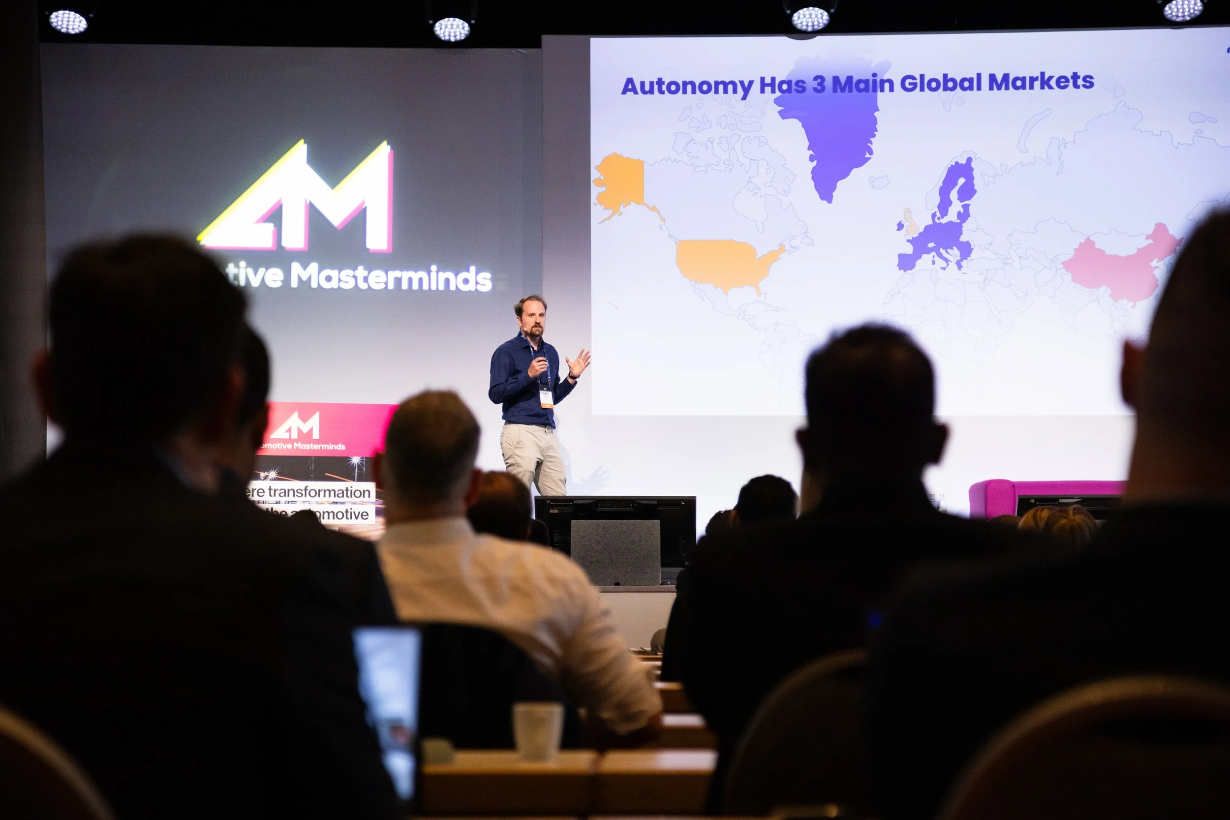 A conference presentation showing a slide titled 'Autonomy Has 3 Main Global Markets' with a world map highlighting regions in different colors. The speaker is a man standing on stage, gesturing with his left hand, in front of an audience.
