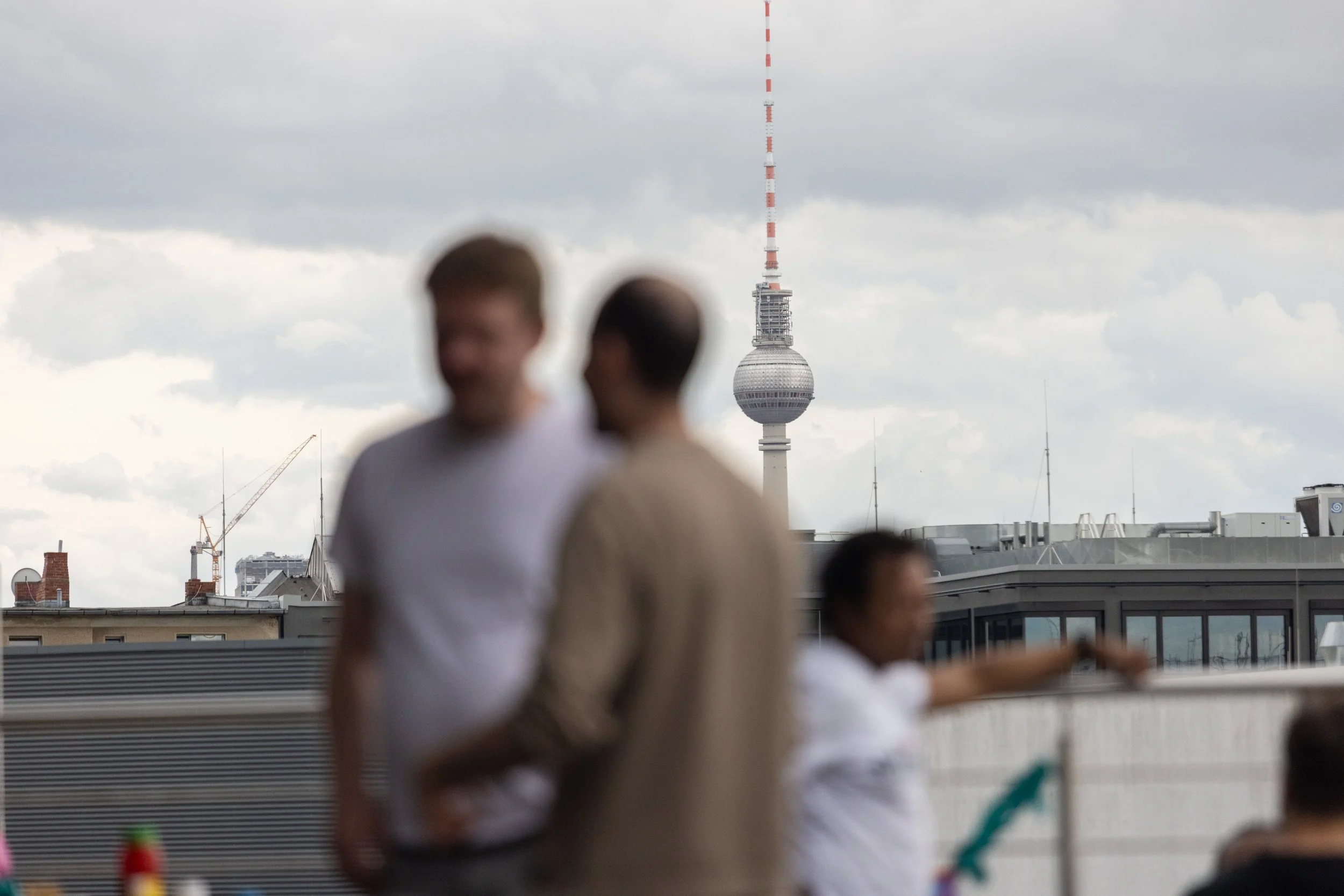 People on a rooftop with blurred faces, with the Berlin TV Tower in the background under cloudy skies.