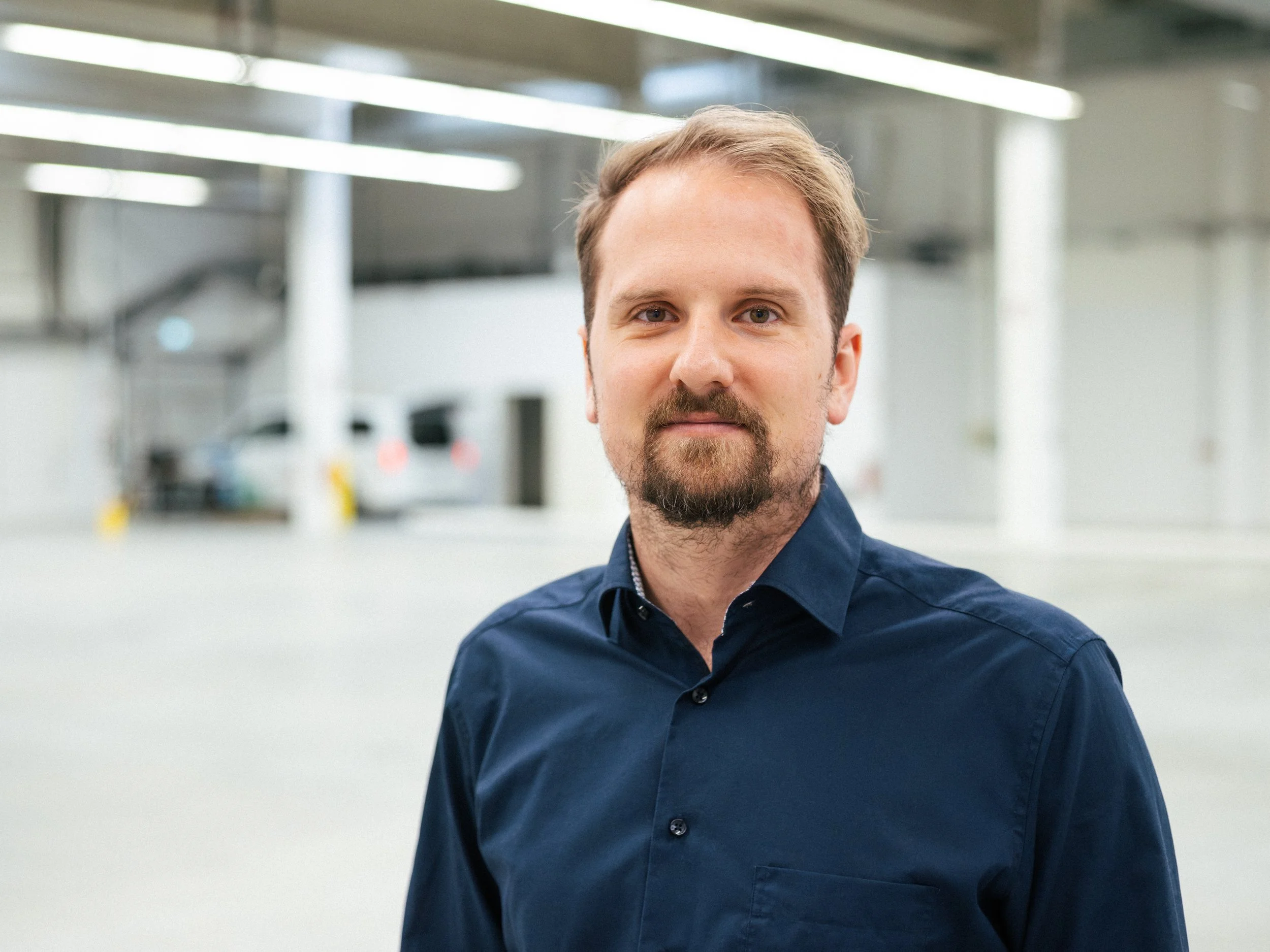 Close-up of a man with blonde hair, beard, and blue eyes, wearing a dark blue button-up shirt, standing in a modern, well-lit indoor space.