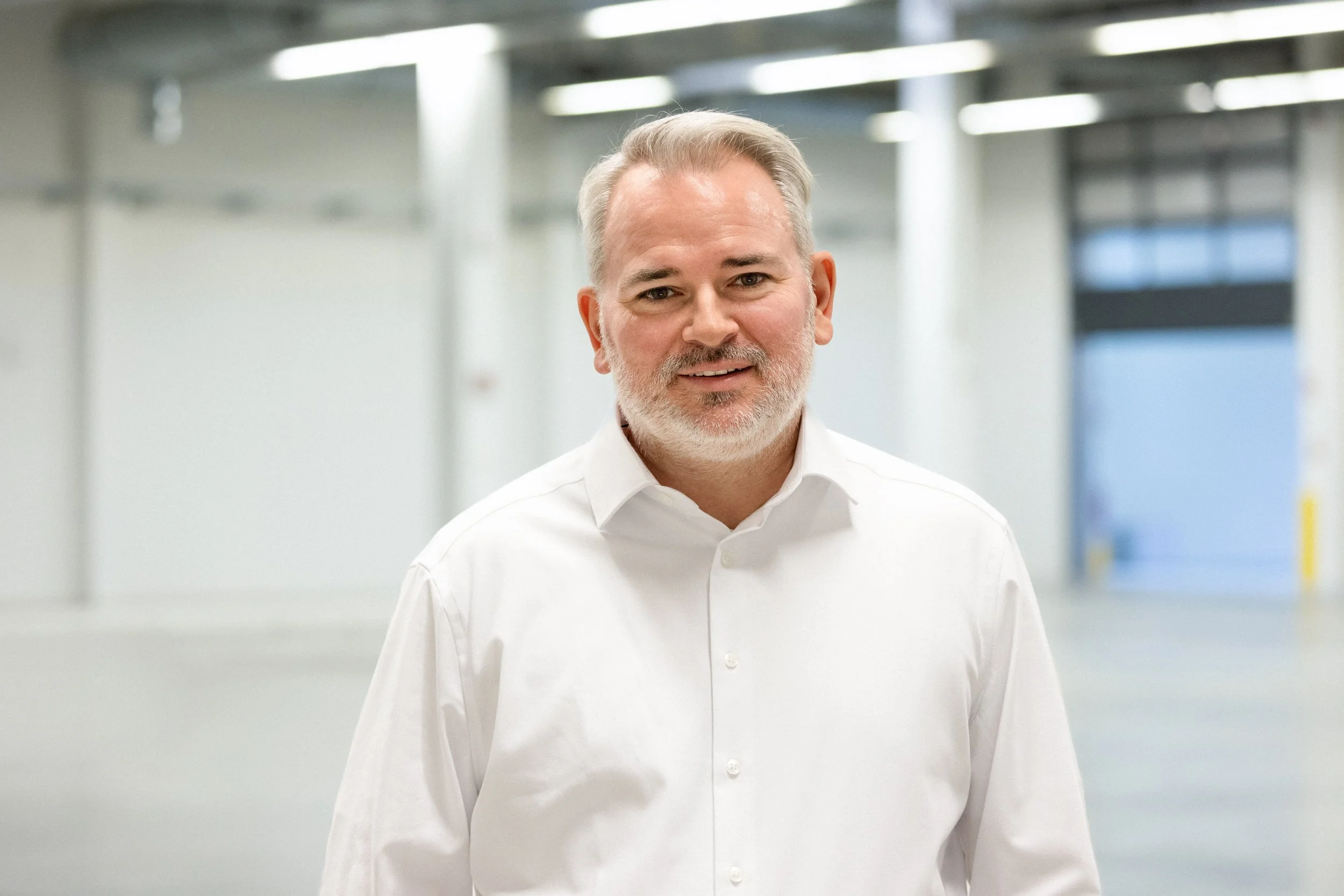 A middle-aged man with gray hair and beard wearing a white dress shirt standing in an indoor setting with bright overhead lighting.