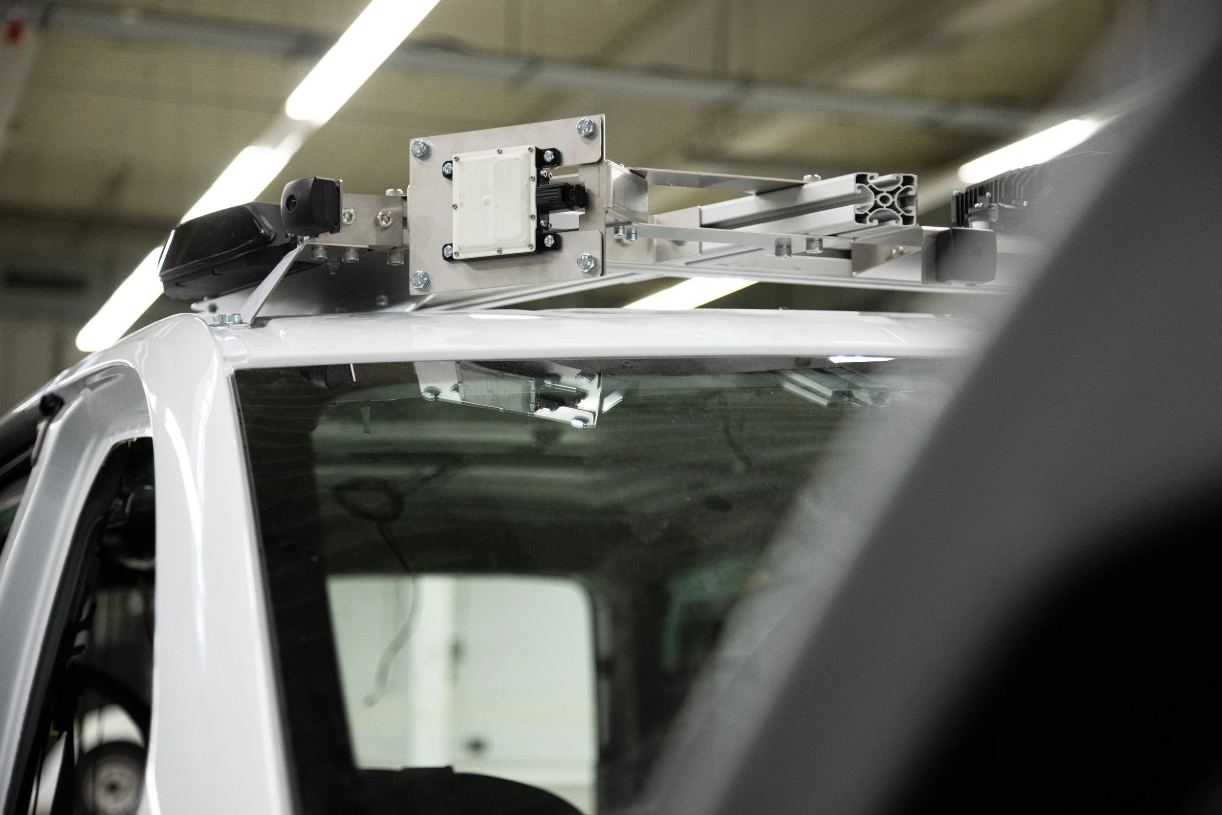 Close-up of a mechanical device mounted on the roof of a white vehicle, with a blurred indoor background and fluorescent lighting.