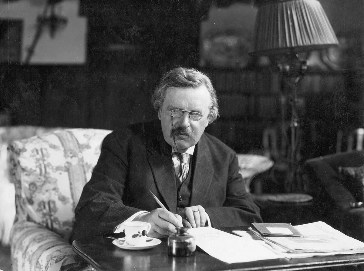 G.K. Chesterton with glasses and a mustache seated at a desk, writing with a fountain pen, surrounded by papers and a teacup, in a dimly lit room.