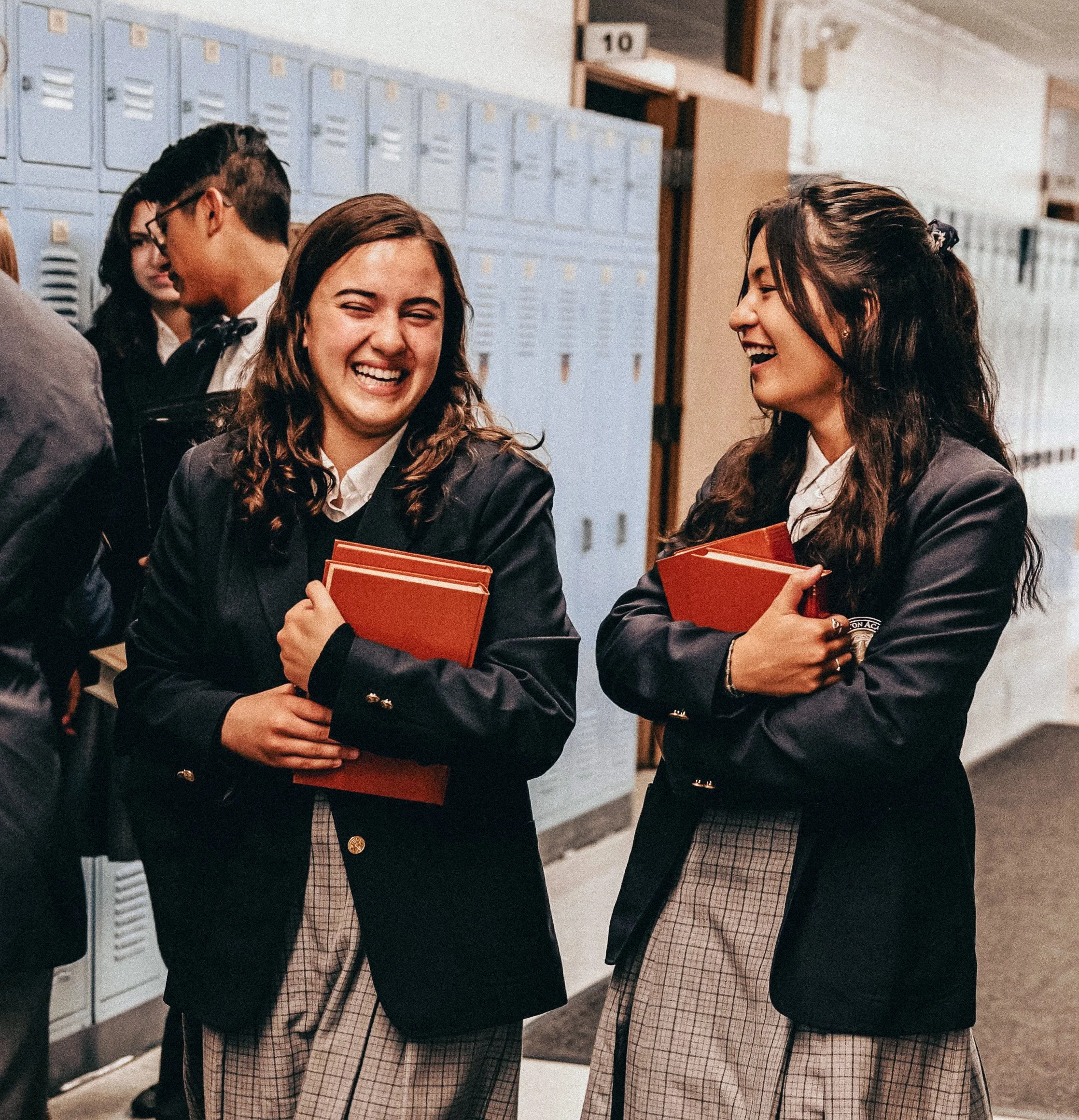 Students in school hallway talking and laughing, holding textbooks, in front of lockers.