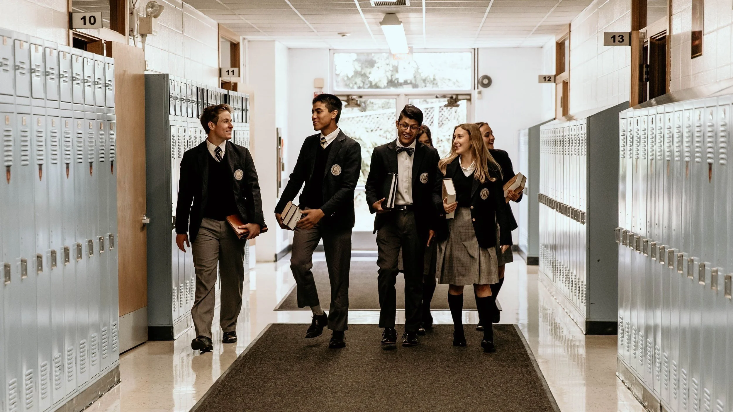 Students walking down a hallway in a school, carrying books and chatting with each other.