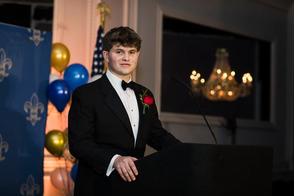 A young man in a tuxedo with a red flower boutonniere speaking at a podium during a formal event, with balloons and a chandelier in the background.