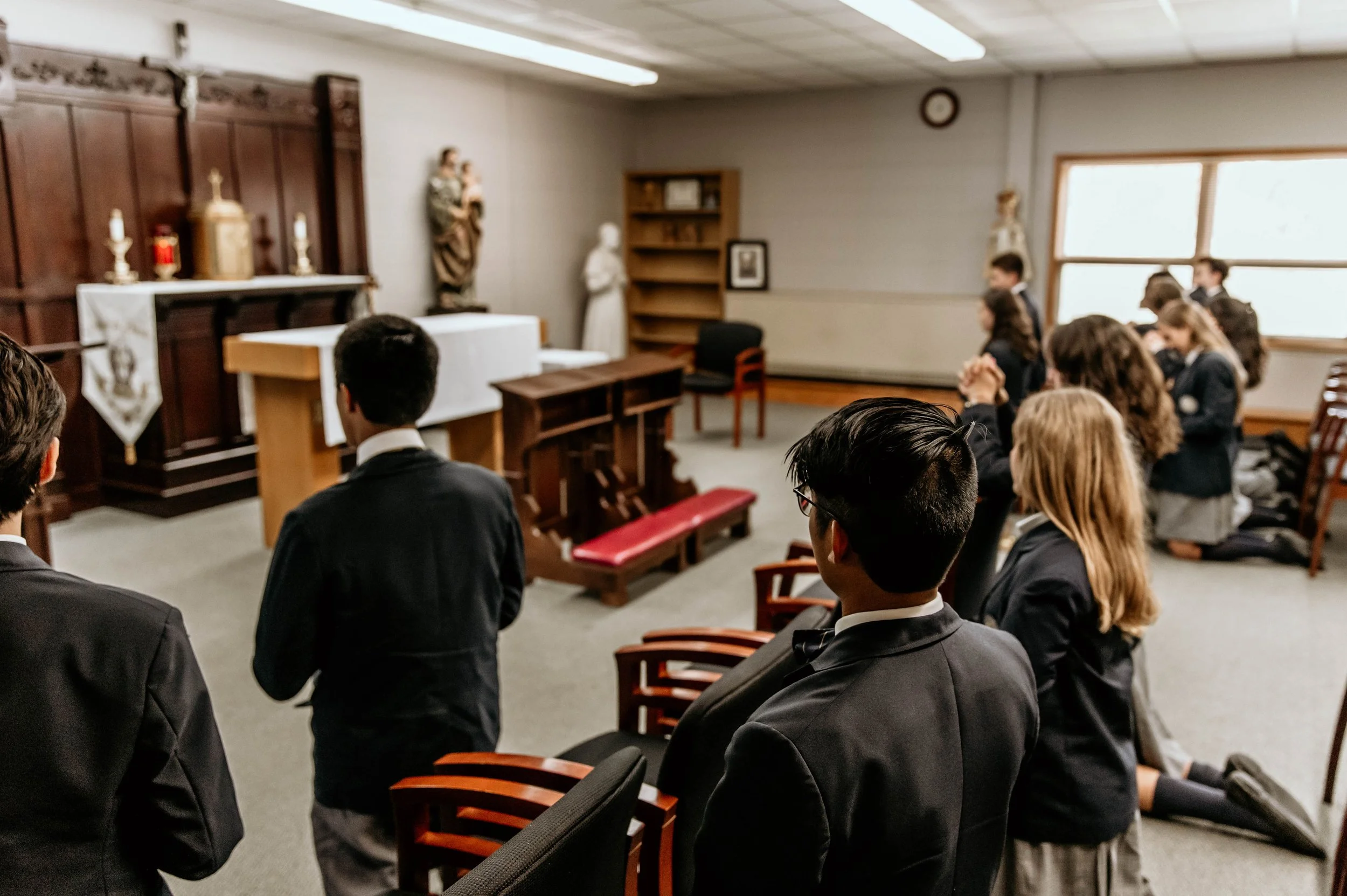Young Chesterton students in uniform kneeling in prayer with hands clasped in a Catholic chapel, surrounded by religious statues.