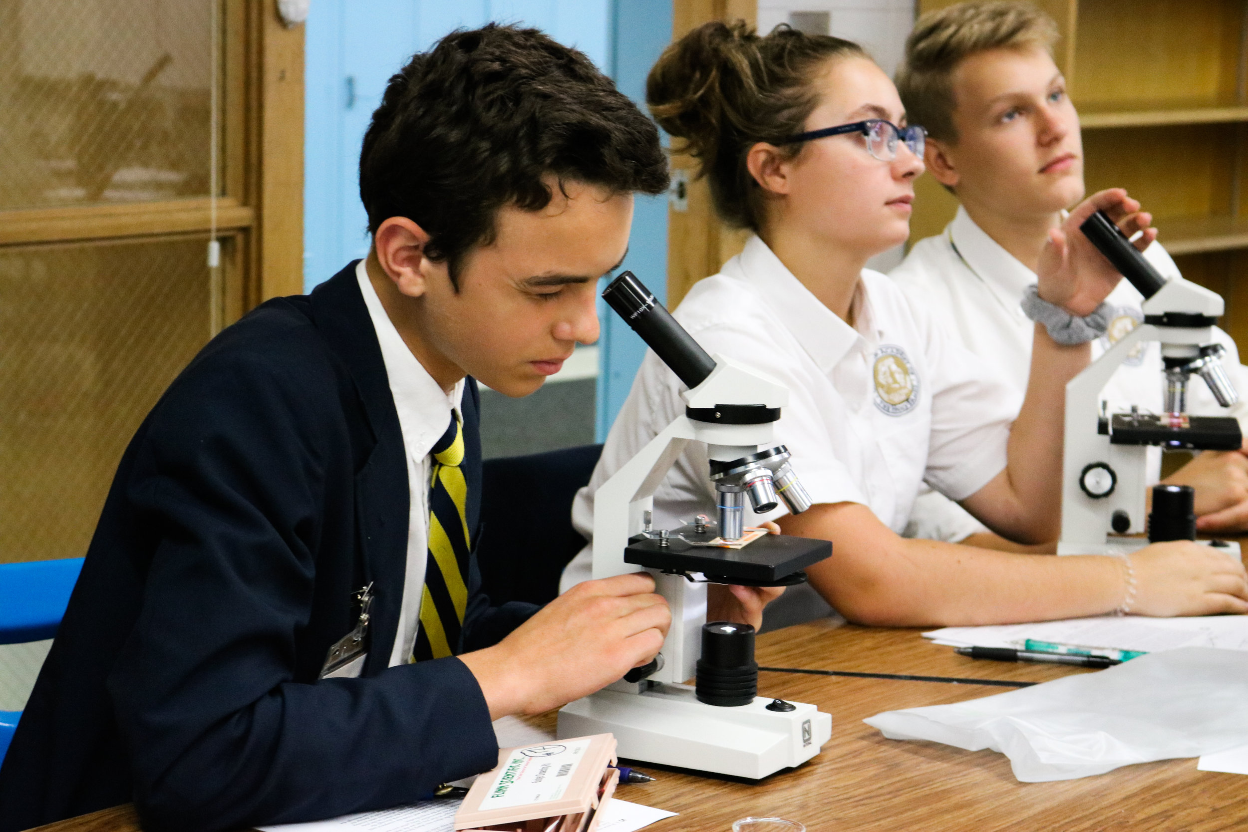 Three students, two boys and one girl, sitting at a table using microscopes in a classroom or laboratory setting.