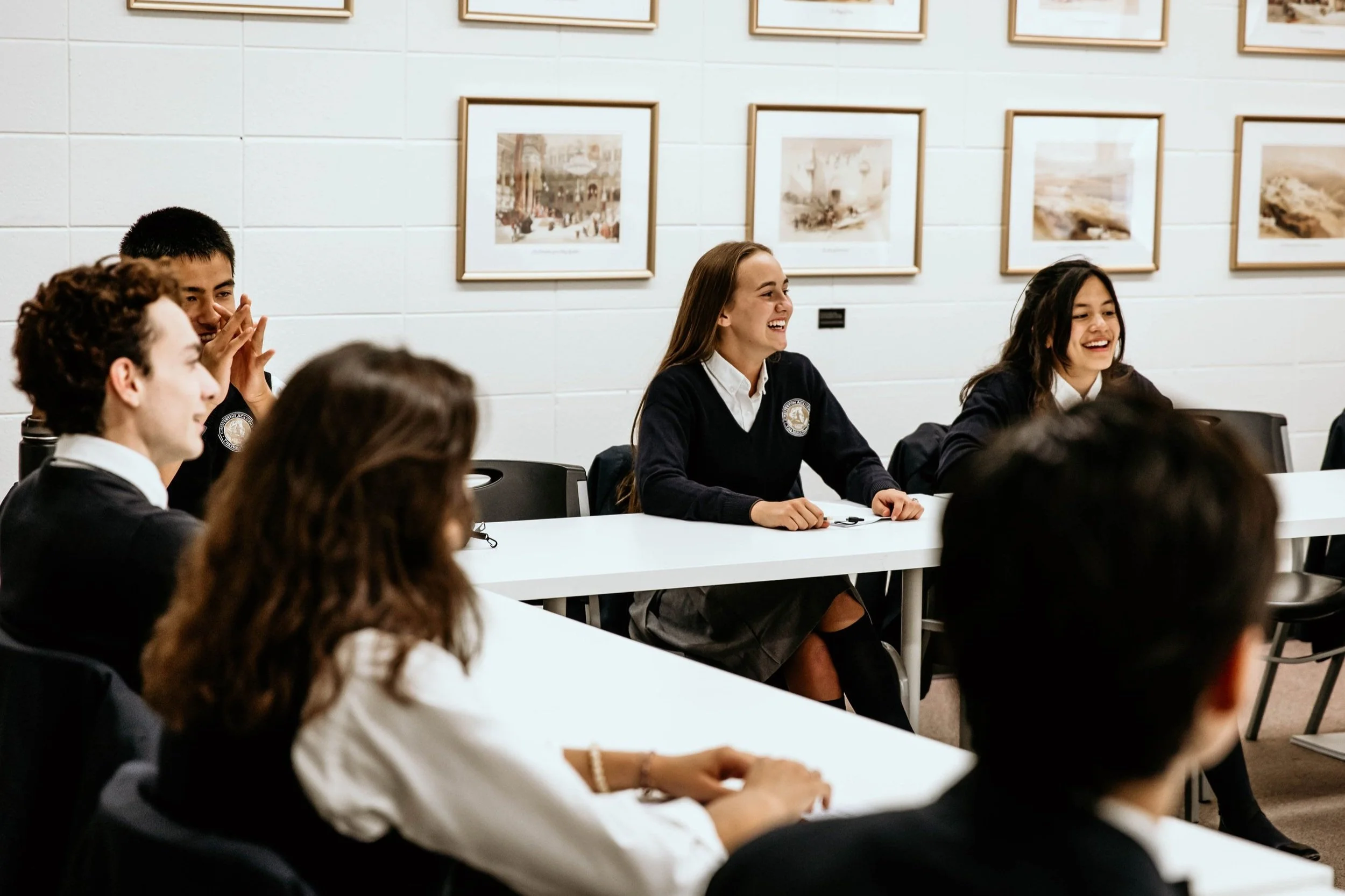 A group of students, mostly girls, sitting at a long table in a classroom, smiling and engaging in a discussion or activity. The classroom has white brick walls decorated with framed artwork.
