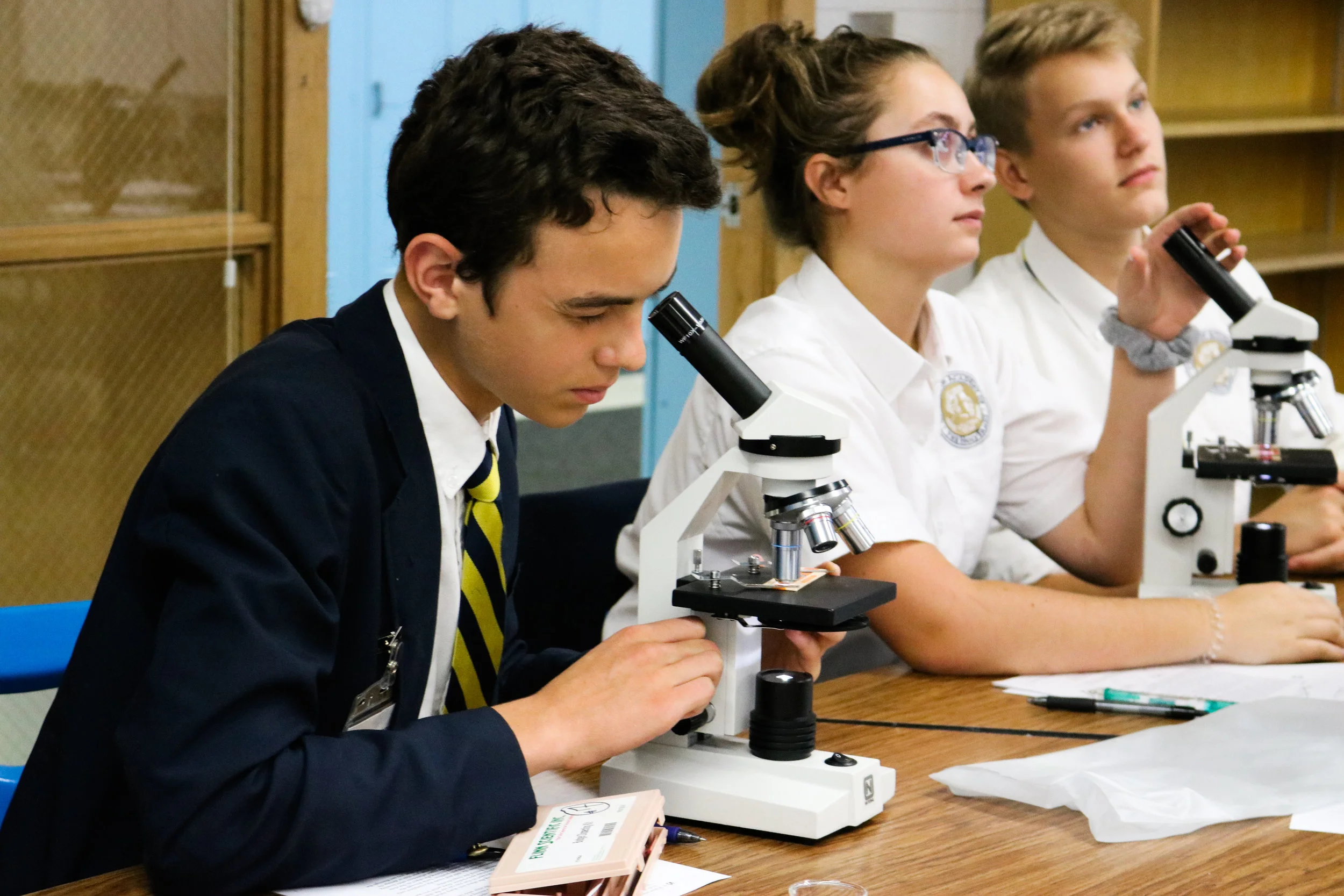 Students in school uniforms using microscopes in a classroom.