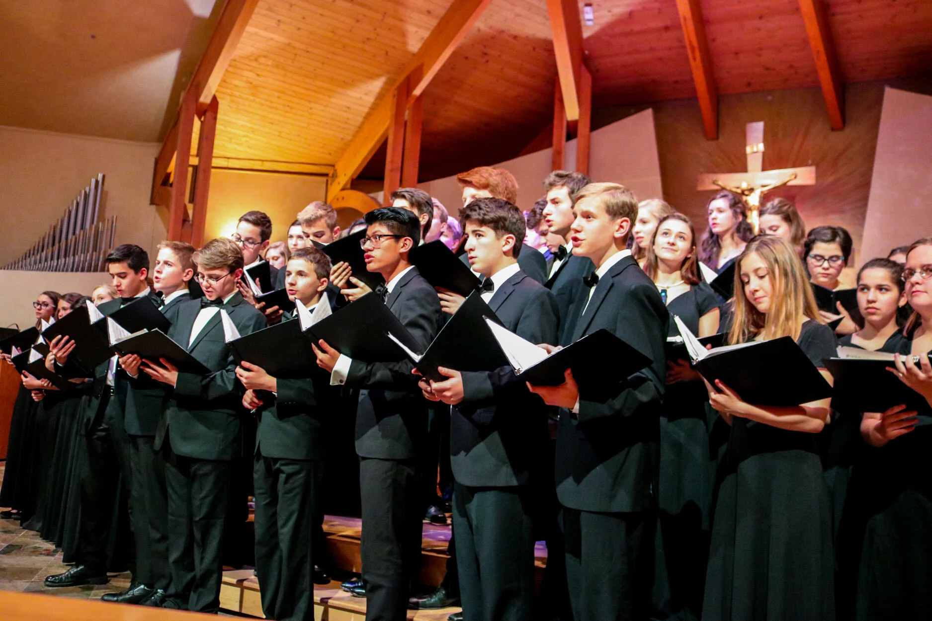 Group of young people singing in choir during a church service, all dressed in formal black attire, with some holding black folders with music, inside a church with wood ceiling and crucifix in the background.