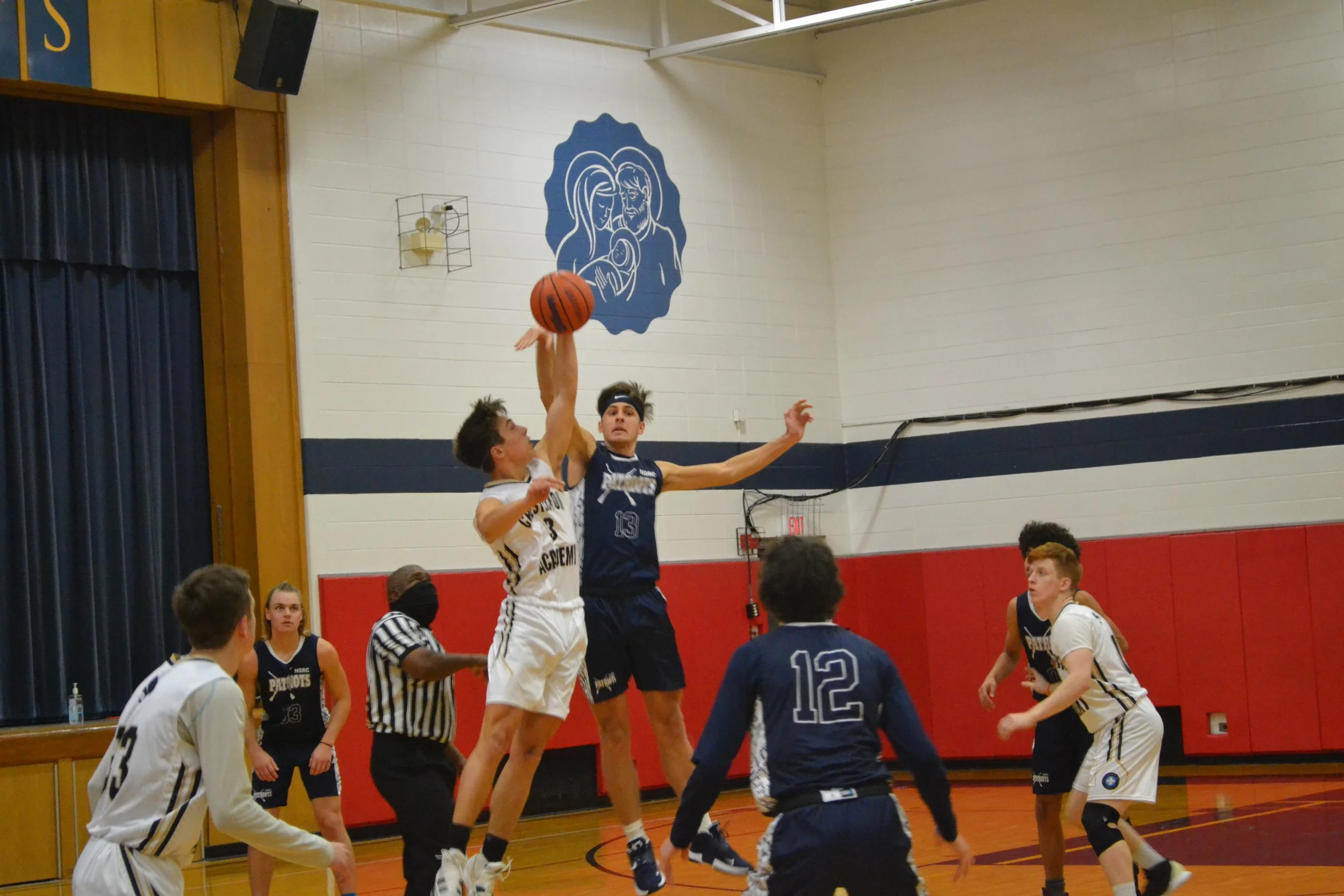 Two basketball players, one in a white jersey and the other in a dark blue jersey, jump to contest a shot near the basket in a gymnasium. Several other players and a referee are visible in the background. A large blue mural of a mother and child is painted on the wall.