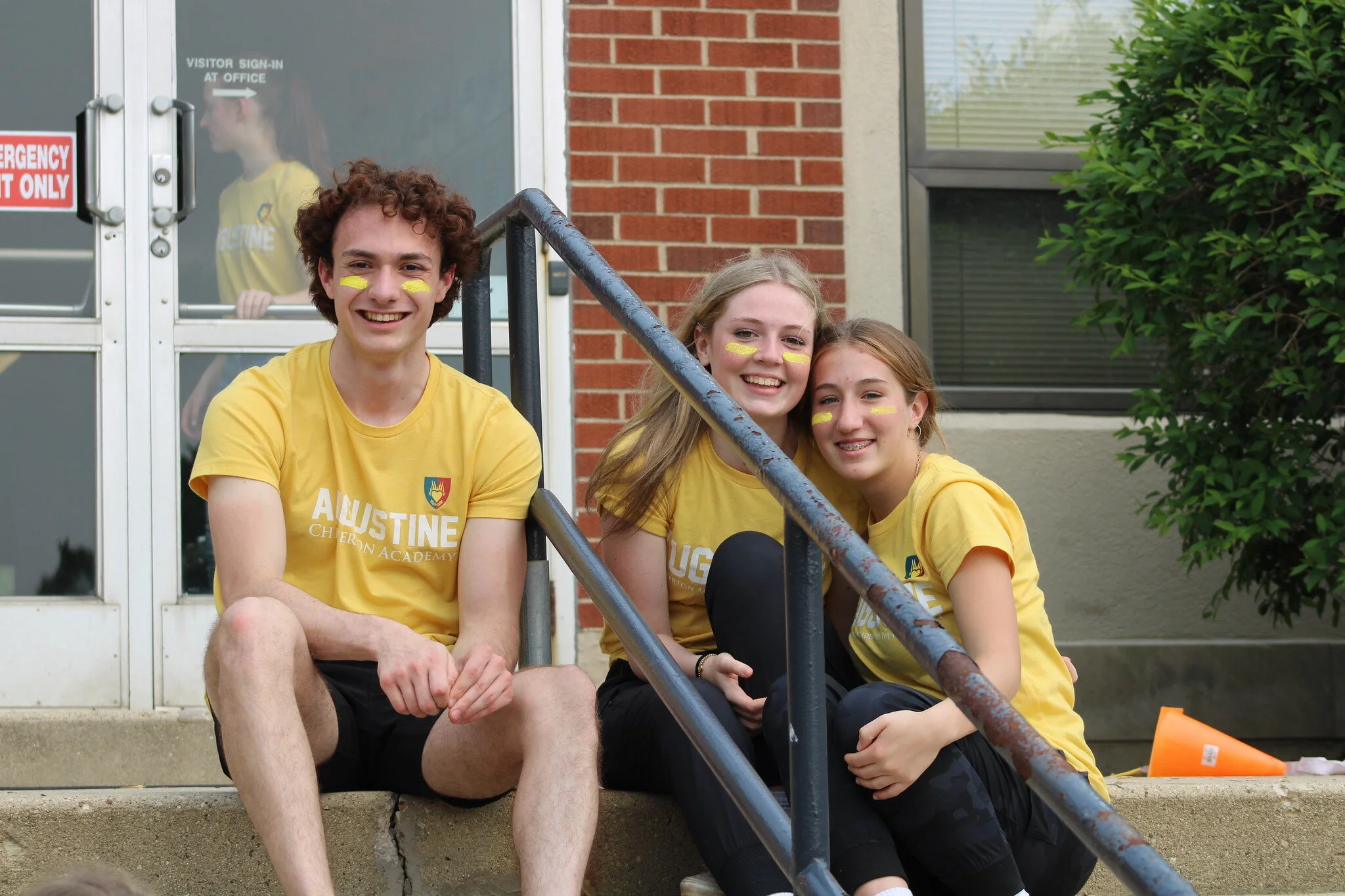 Three students in yellow T-shirts sitting on steps outside a building with a brick wall, window, and green shrubbery, smiling at the camera.