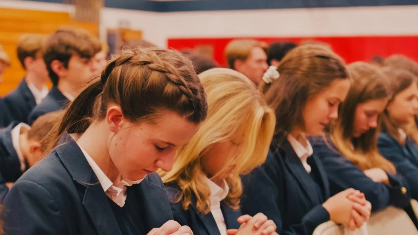 Students in uniform praying with their hands clasped in a gym.
