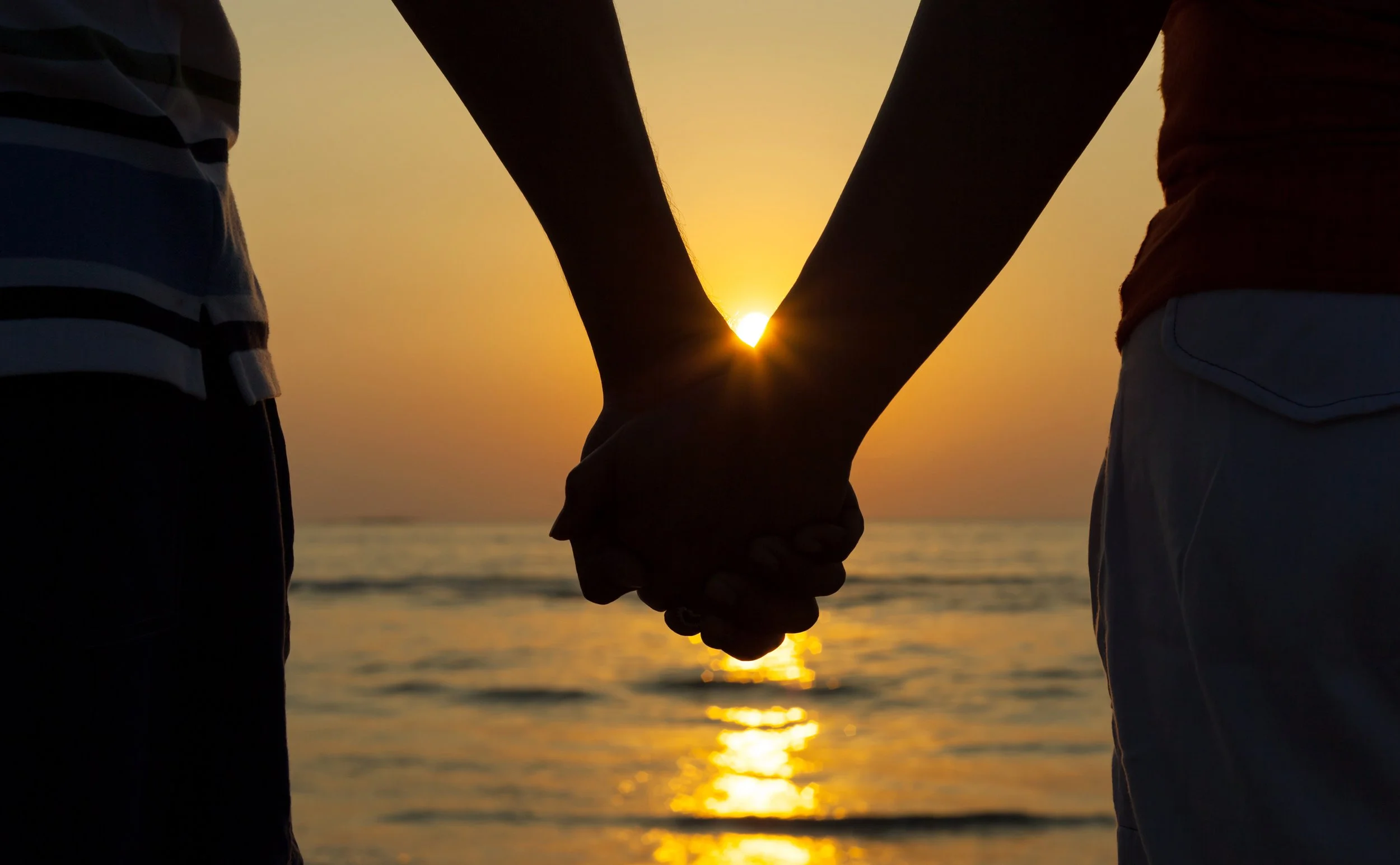 Hands holding in front of a sunset on the beach.