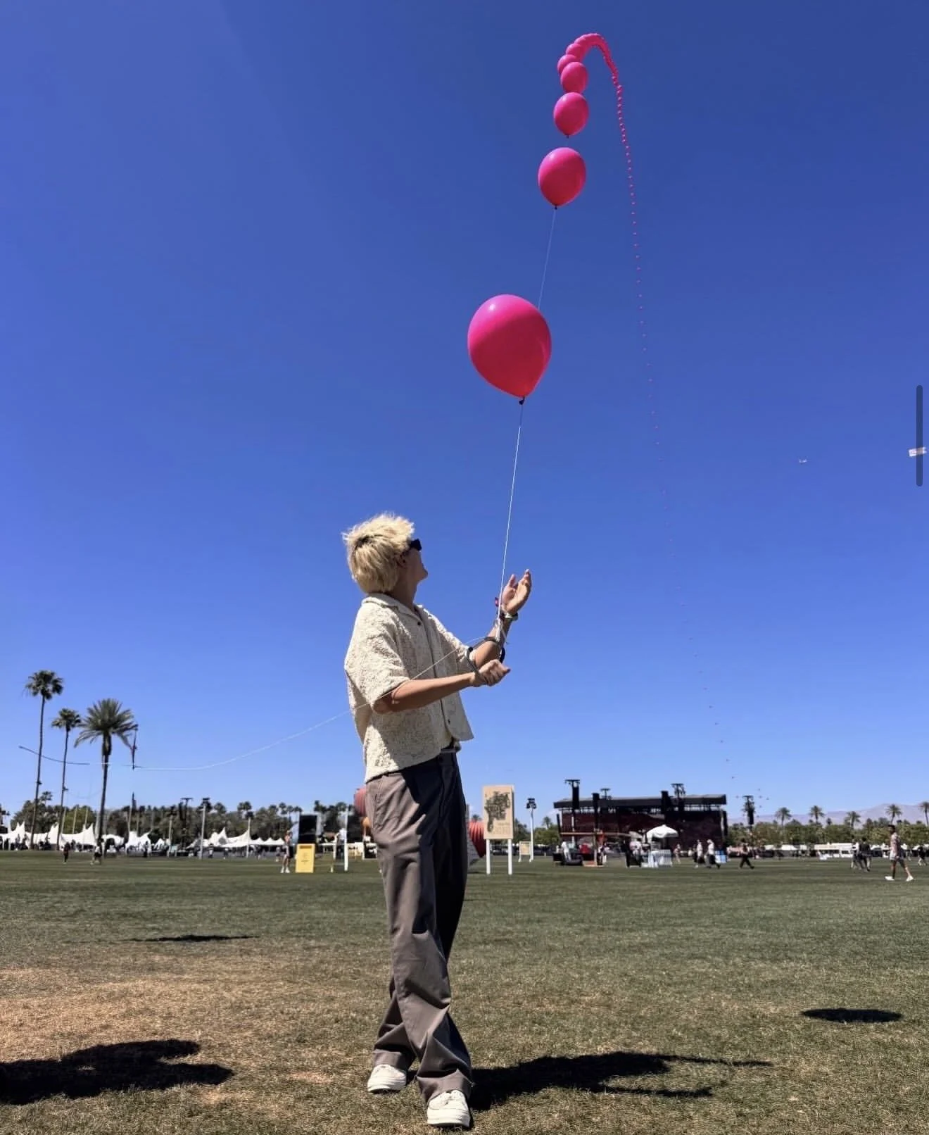 A person standing on a grassy field holding a string attached to five pink balloons floating in a clear blue sky. There are palm trees and a stage in the background.