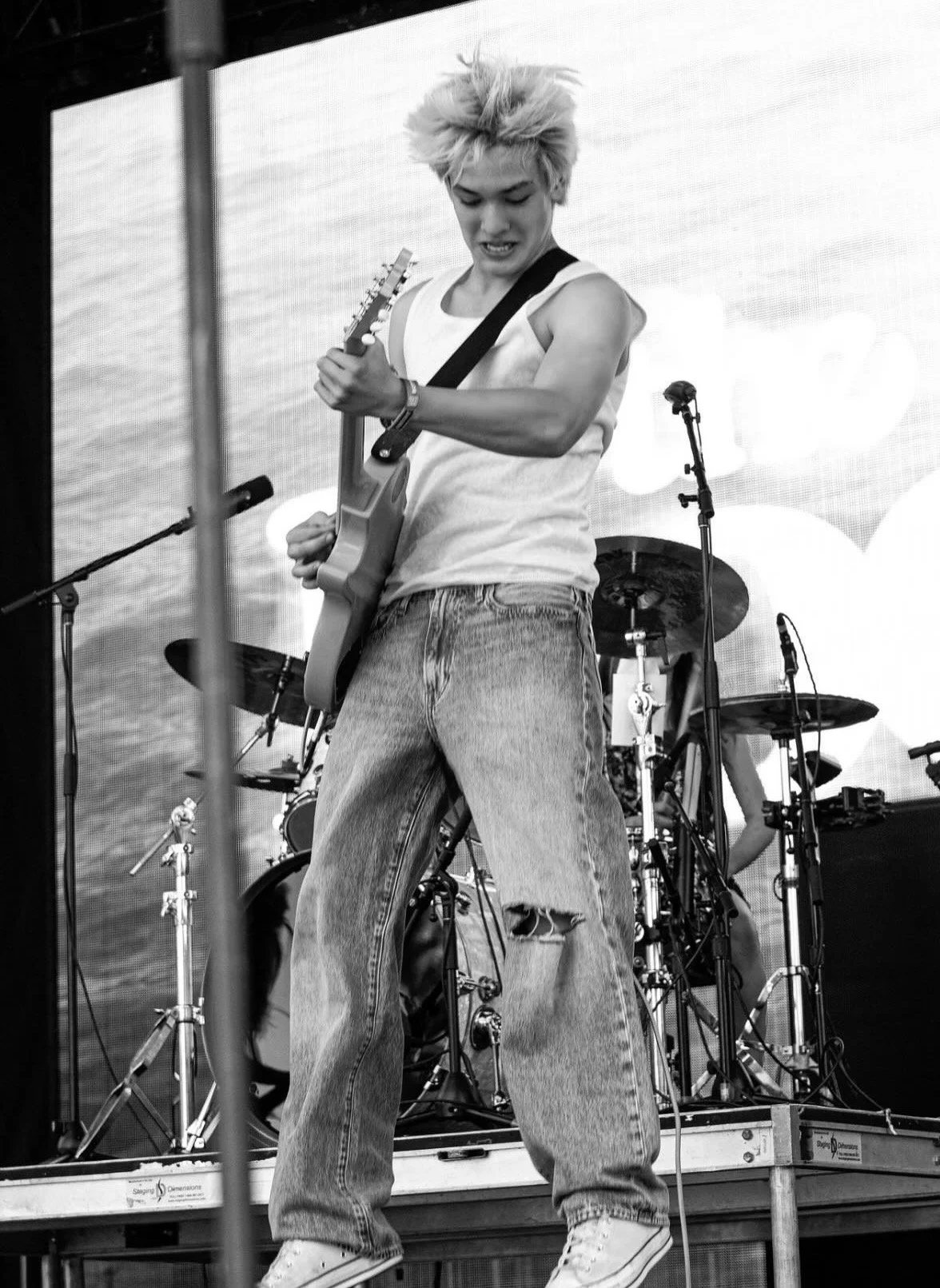 A young woman with spiky blonde hair playing an electric guitar on stage with a drum set in the background