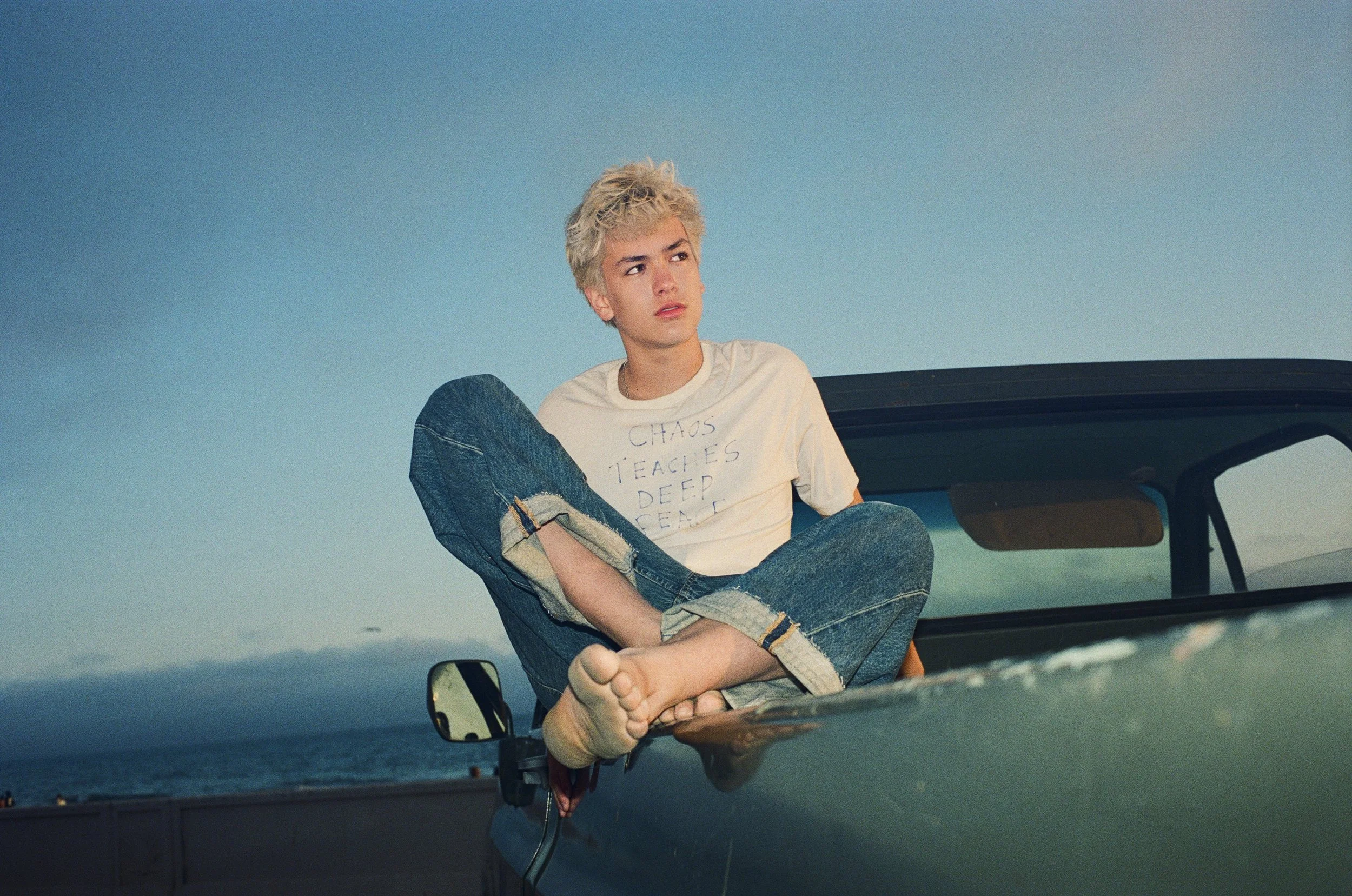 A young man with curly blonde hair sitting on the hood of a car with the ocean in the background.