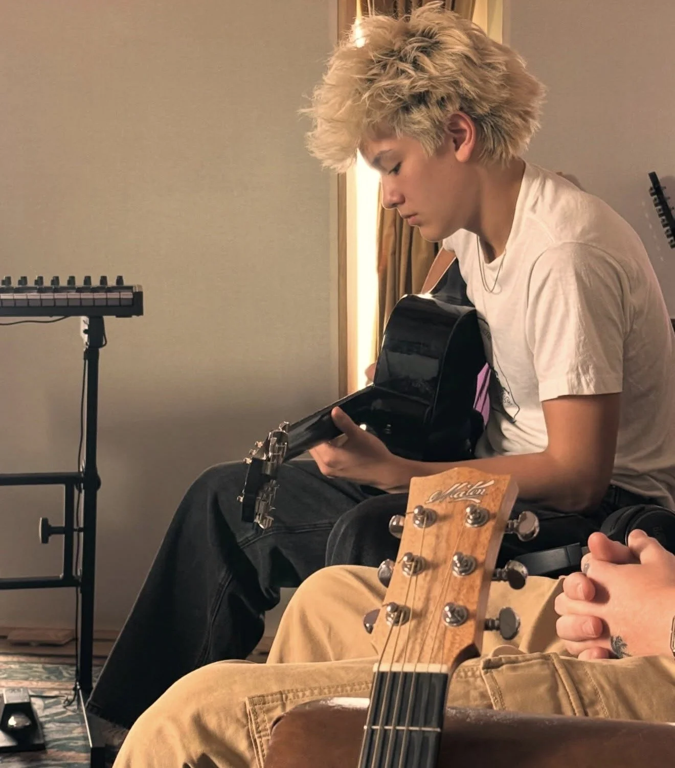 A young person with curly blonde hair sitting in a room, looking into a black cases with a guitar inside, surrounded by musical equipment.