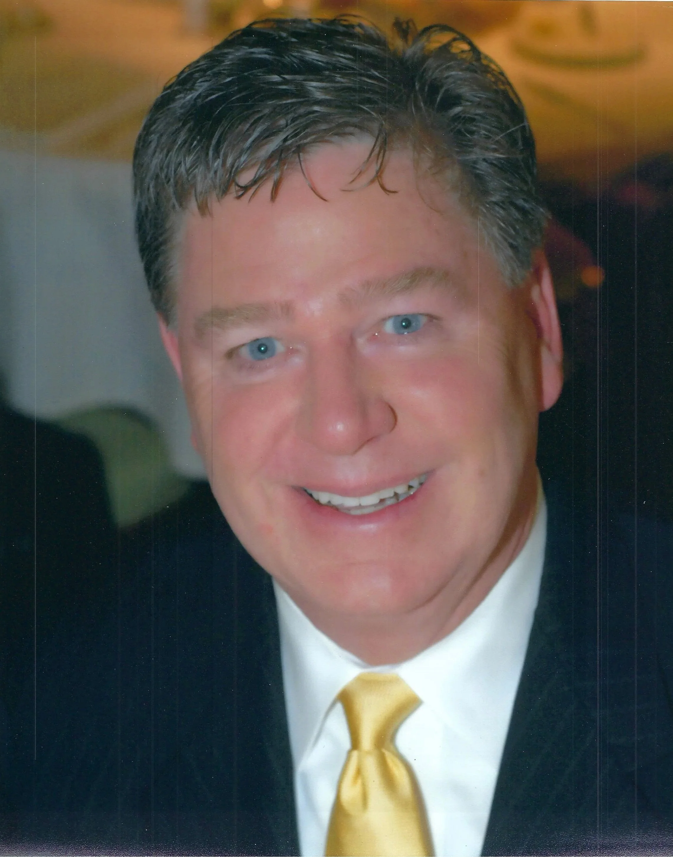 A man with short, dark hair, blue eyes, and light skin, dressed in a suit, white shirt, and yellow tie, smiling in a professional portrait.