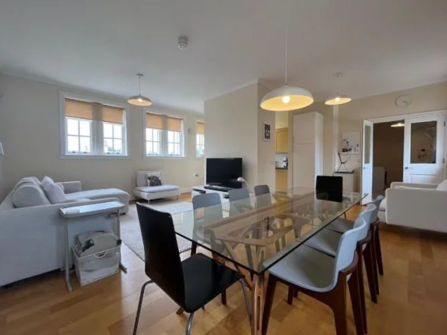 Open-plan living and dining room with a glass dining table, six chairs, white and beige sofas, a TV, and large windows with beige blinds.
